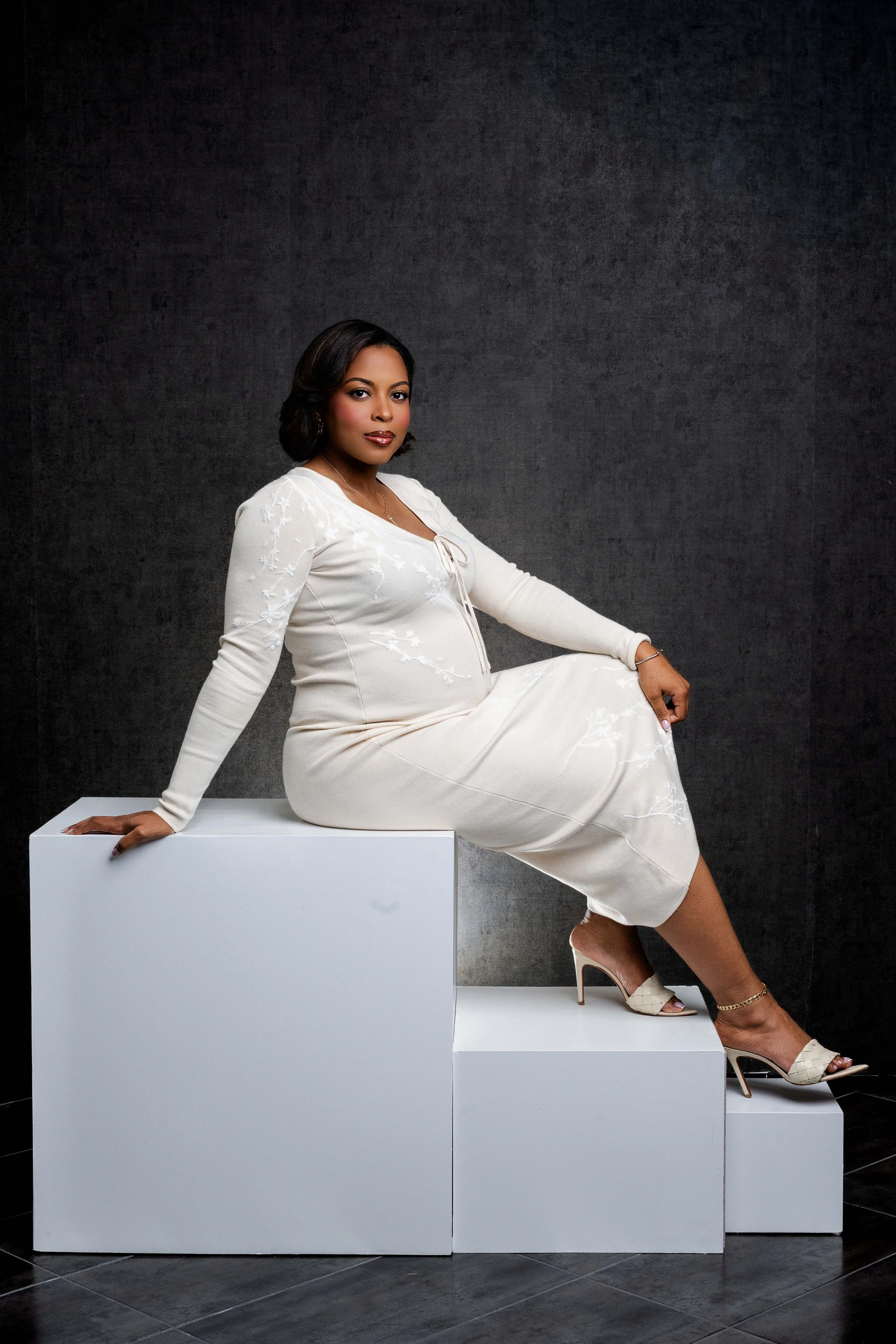 A woman in a white dress and high heels sits on white geometric blocks against a dark background.