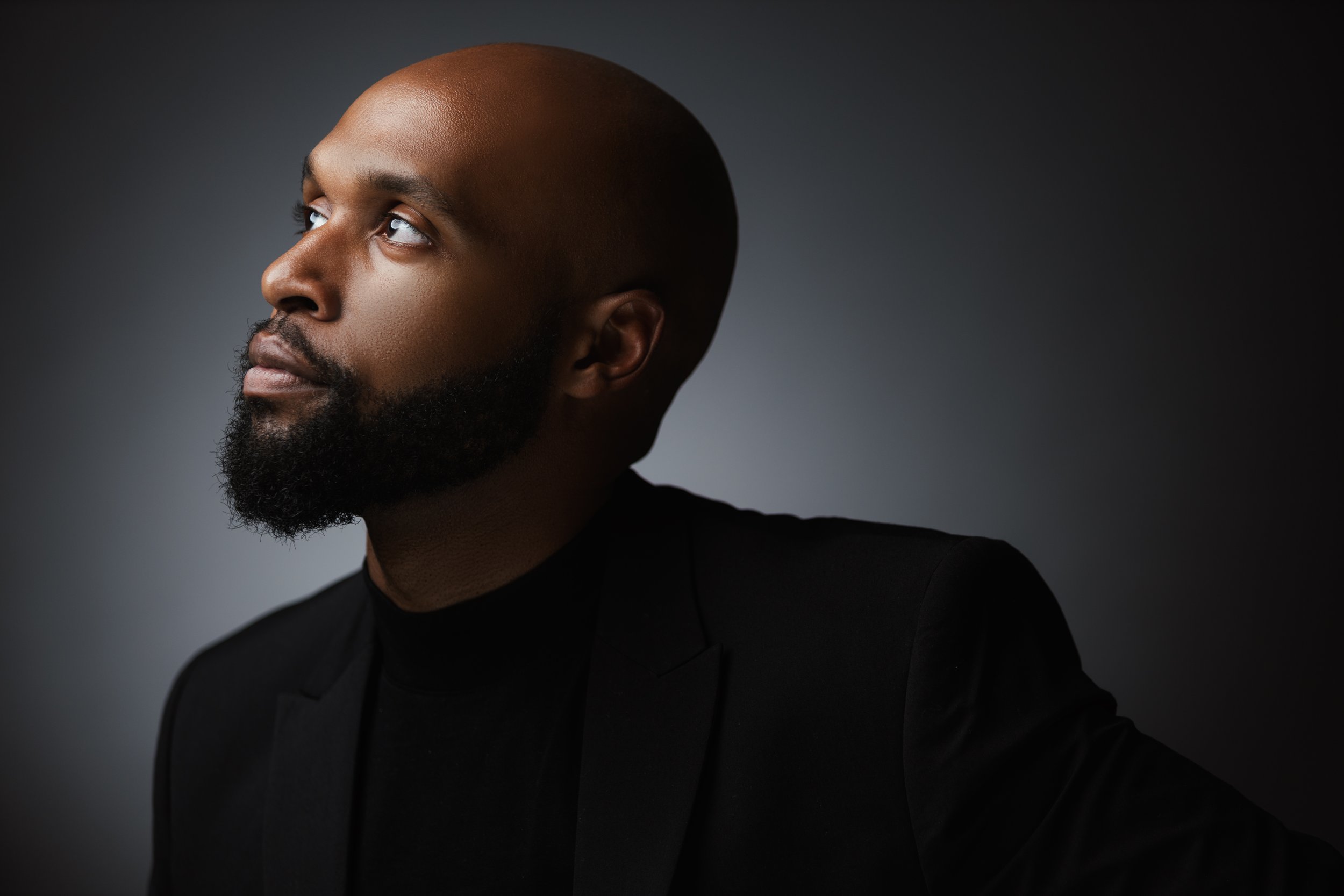 A portrait of a man in a black suit against a dark gray background, looking slightly upward to the left with a contemplative expression.