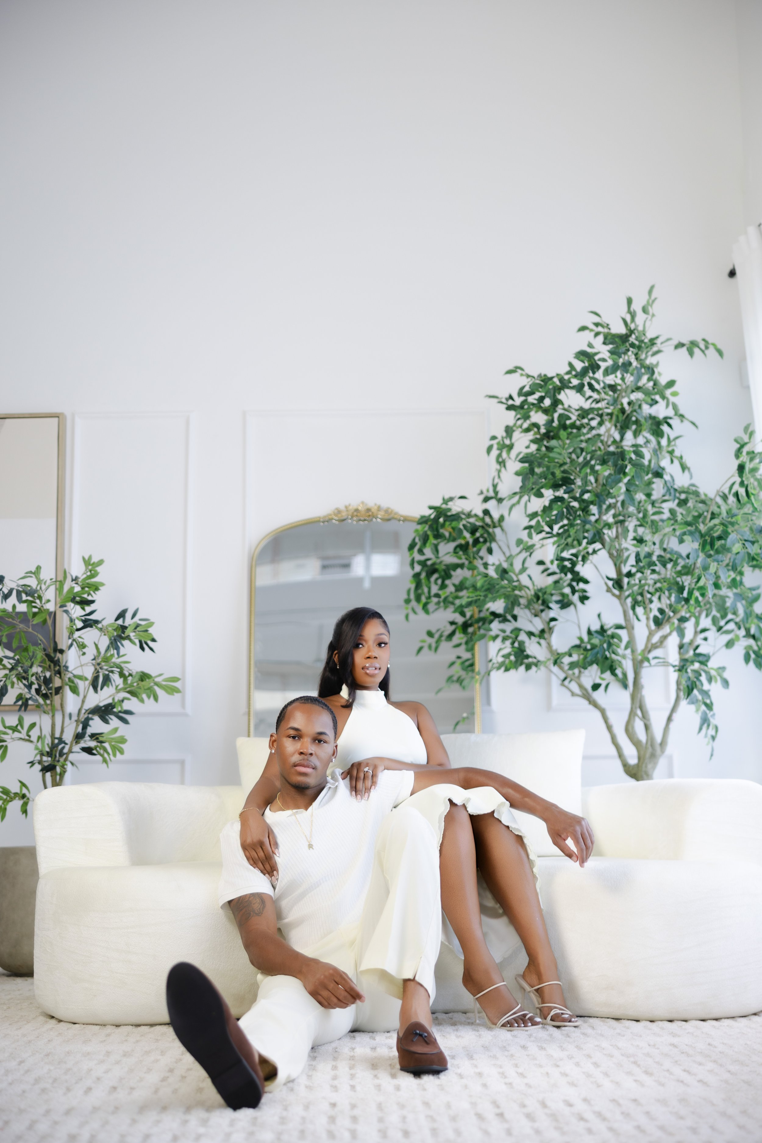 A man lying on a white sofa with a woman sitting behind him in a bright, modern room with large potted plants, white walls, a mirror, and a white rug.