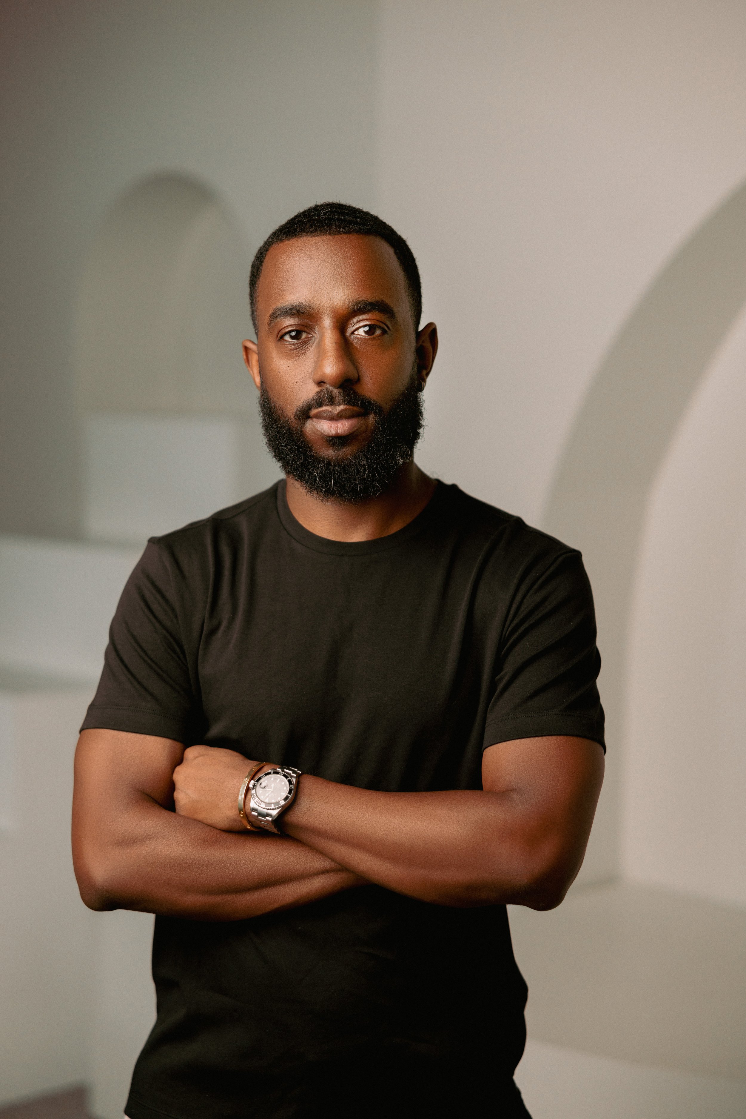 A young man with dark hair, a full beard, and medium skin tone is standing indoors with crossed arms, wearing a black T-shirt and a silver watch on his left wrist. He looks confidently at the camera with a neutral expression against a neutral backgro