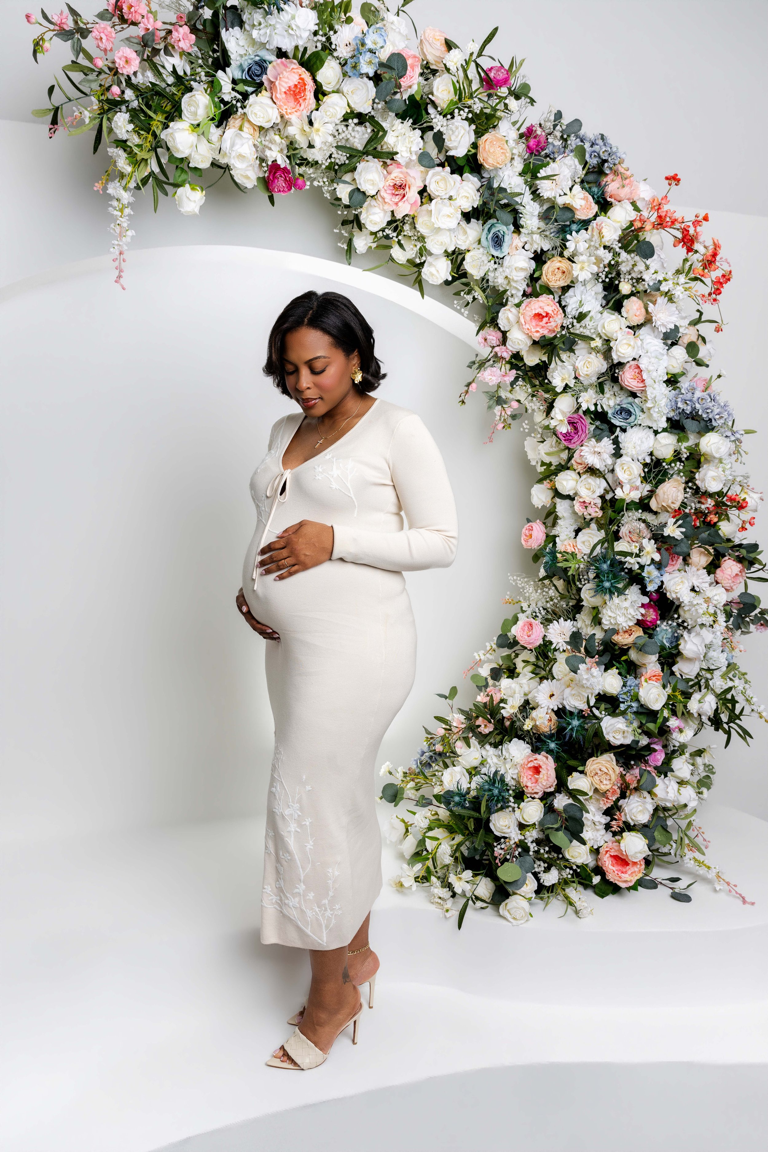 A pregnant woman in a long white dress with floral embroidery standing in front of an arch made of white and pastel-colored flowers including roses, peonies, and hydrangeas, against a white backdrop.