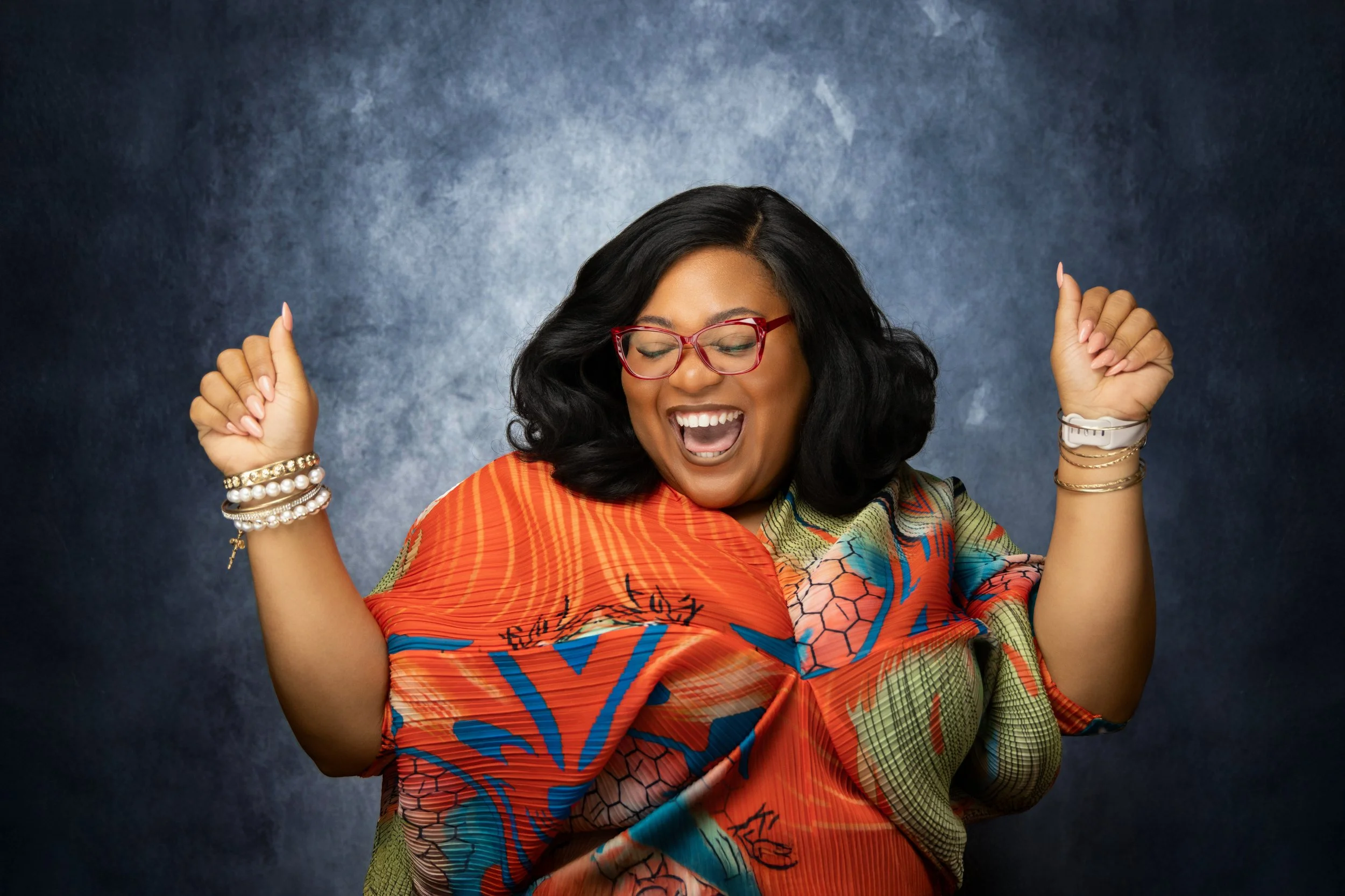 A smiling woman with black hair, wearing red glasses, a colorful patterned shirt, and jewelry, dancing with her eyes closed against a dark textured background.