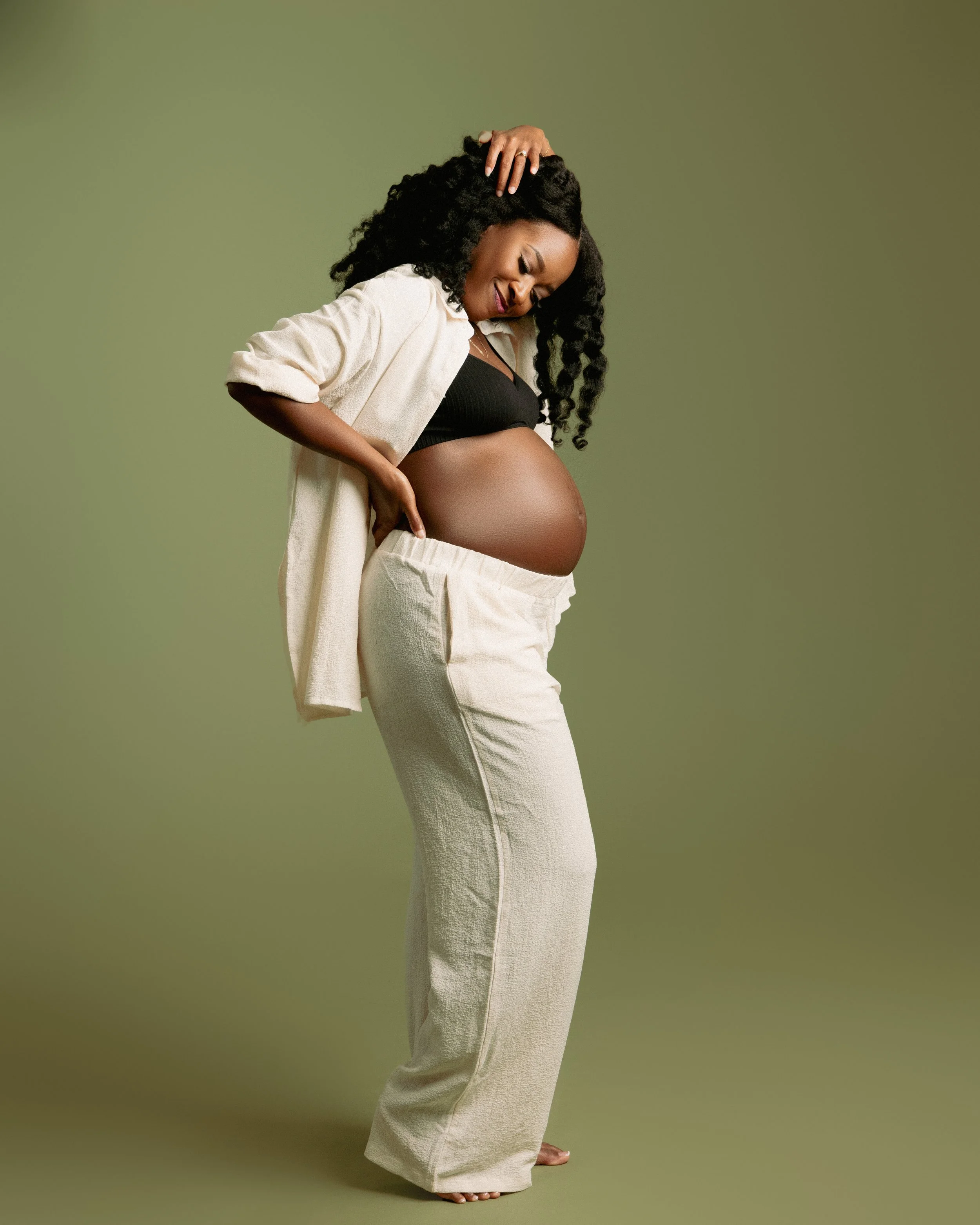 A pregnant woman with curly black hair smiling, looking down, wearing a black top, beige robe, and beige pants, standing against a solid green background.