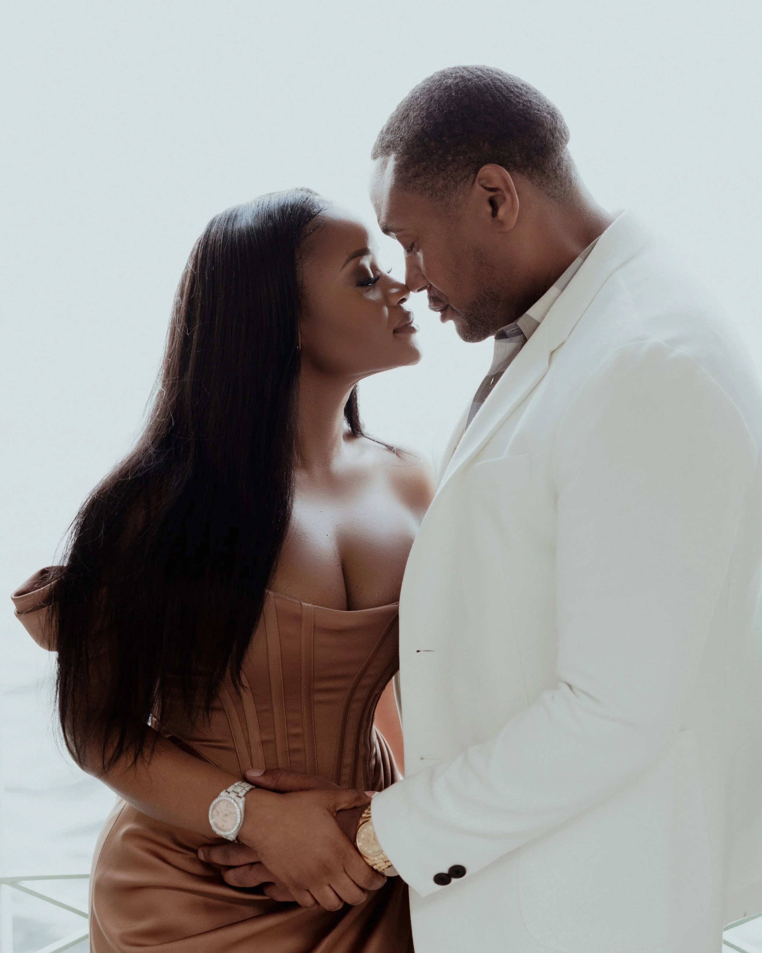 A couple standing close together with foreheads touching and eyes closed, holding hands, against a plain light background.