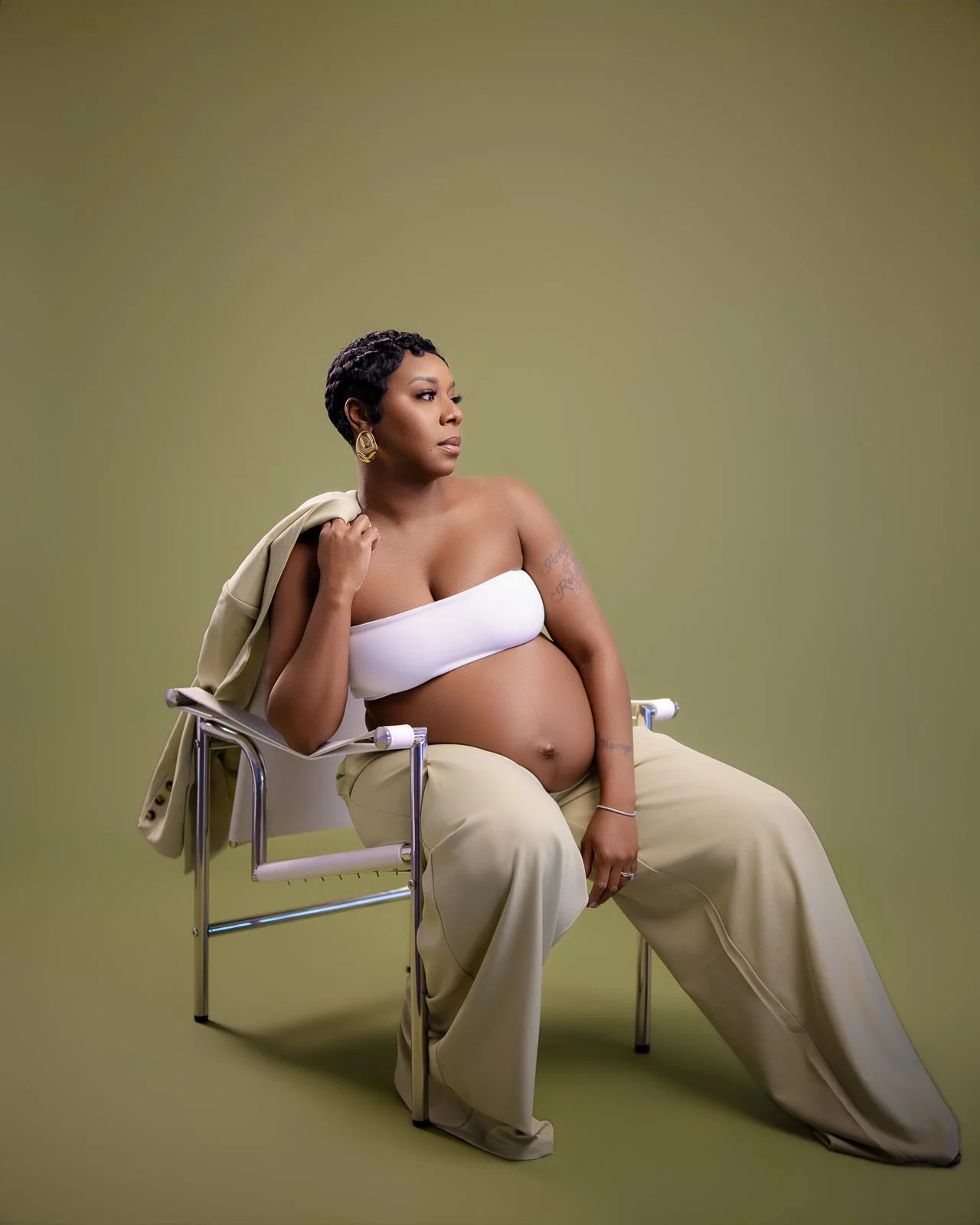 A pregnant woman sitting on a chair, wearing a white bandeau top and beige pants, with a beige blazer draped over her shoulder, against a plain green background.