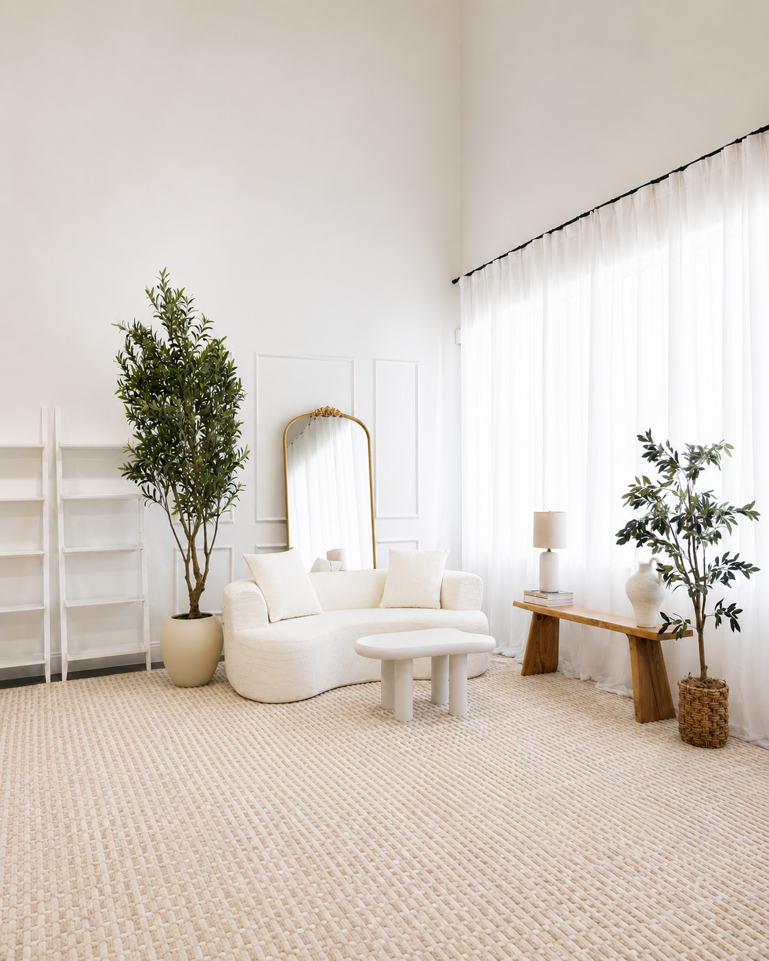 Bright living room with white sofa, two large potted plants, a wooden bench with a table lamp and vase, a tall mirror, and sheer white curtains.