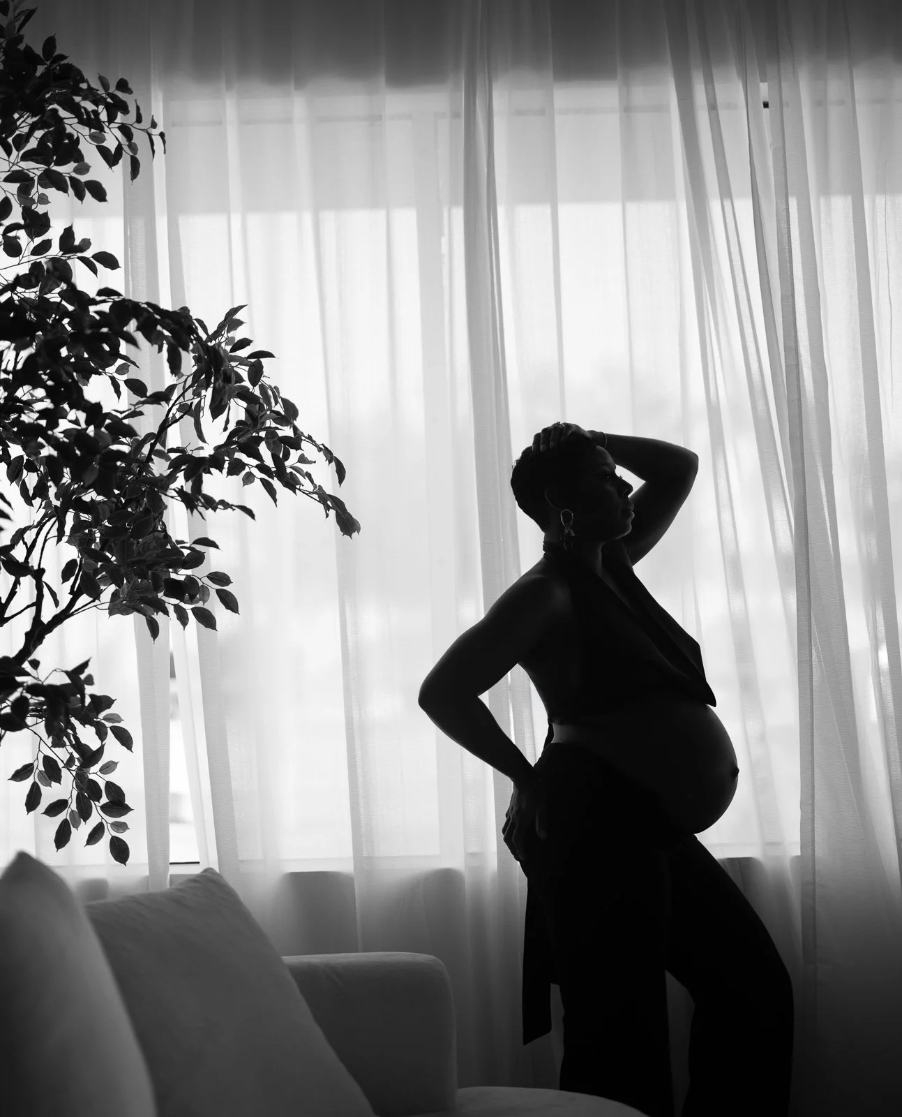 Silhouette of a pregnant woman standing by a window with sheer curtains, with a plant nearby, in black and white.