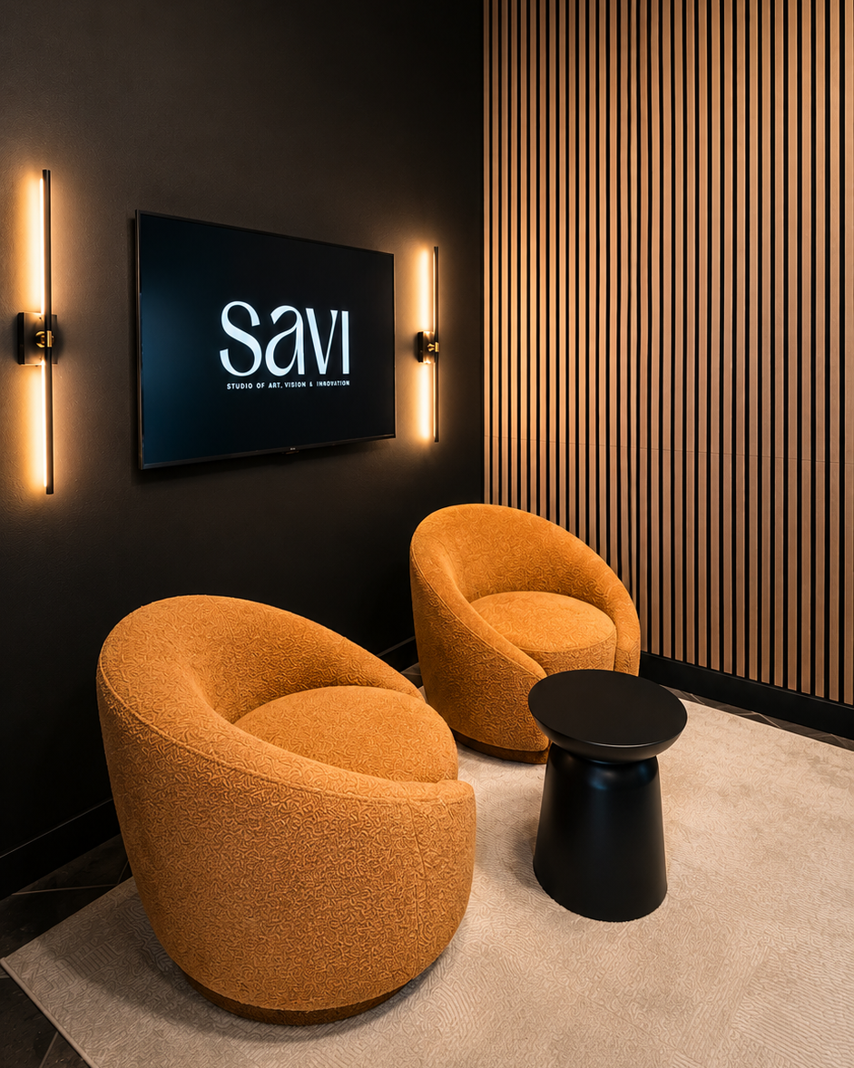 A modern lounge area with two orange upholstered chairs, a small black side table, a wall-mounted television displaying 'Savi,' and stylish wall lighting on a dark wall with wooden paneling.