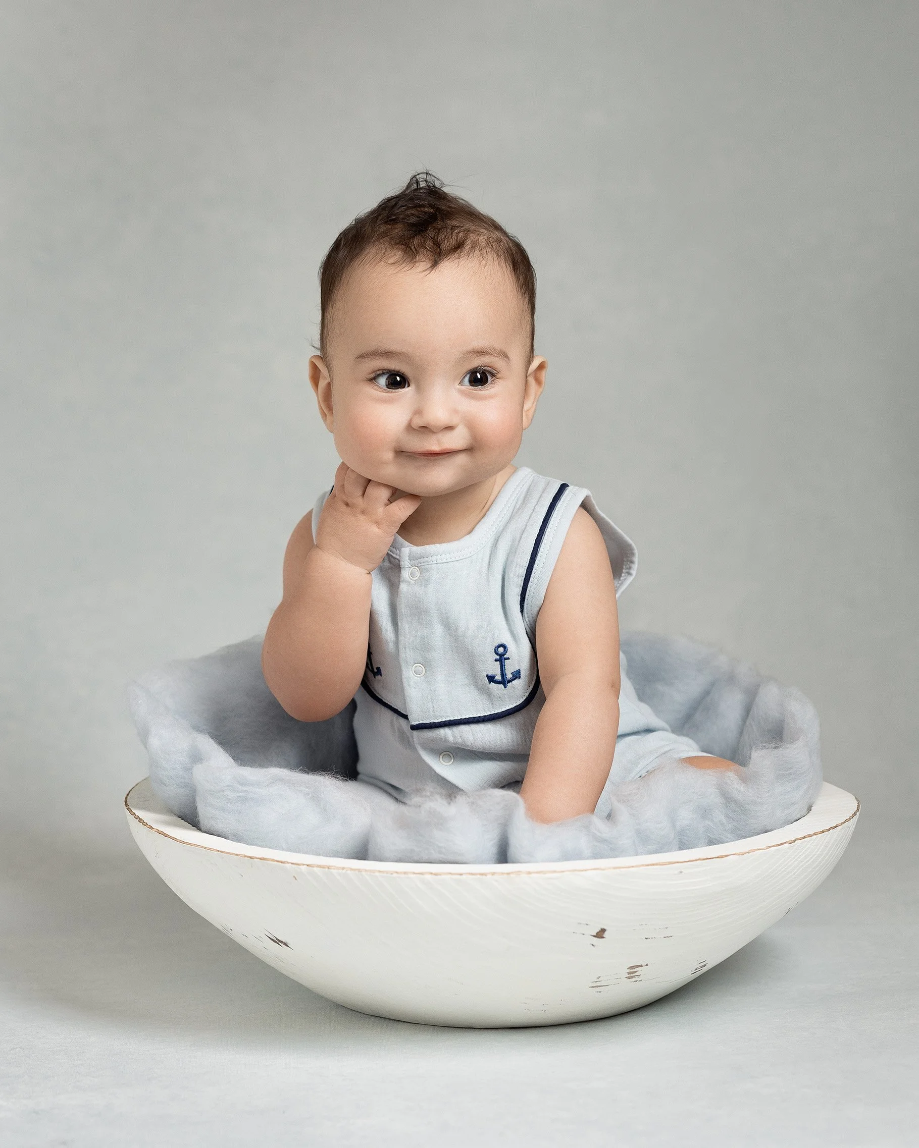 A baby with short brown hair sitting inside a shallow white bowl, with a gray cushion lining the interior, against a plain light gray background.