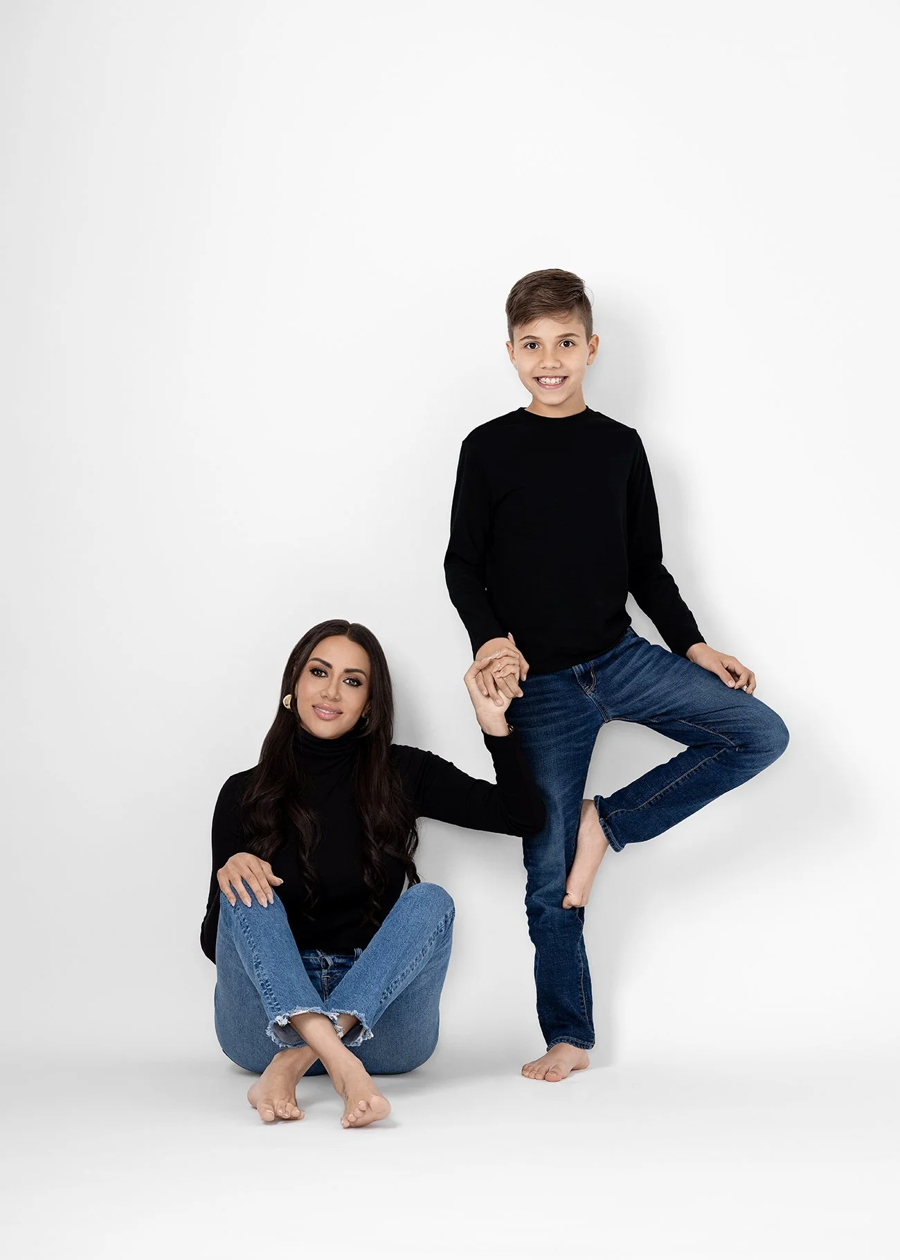 A woman sitting on the floor with her legs crossed and a young boy standing beside her, holding her hand. They are both wearing black tops and blue jeans, and are posed against a plain white background.
