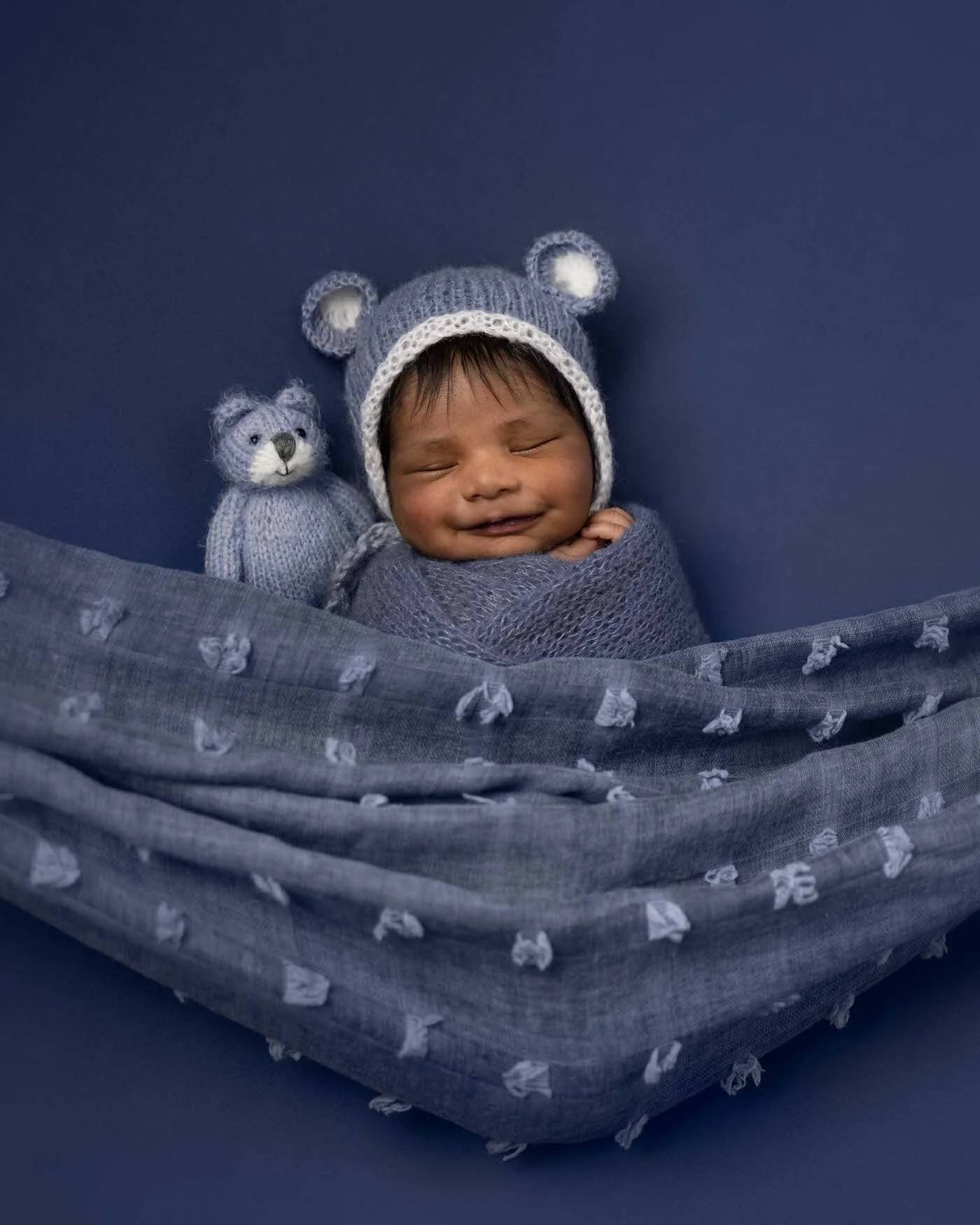 Smiling baby boy wrapped in a gray knitted blanket, wearing a gray bear hat, sitting against a blue backdrop with a matching gray knitted teddy bear.