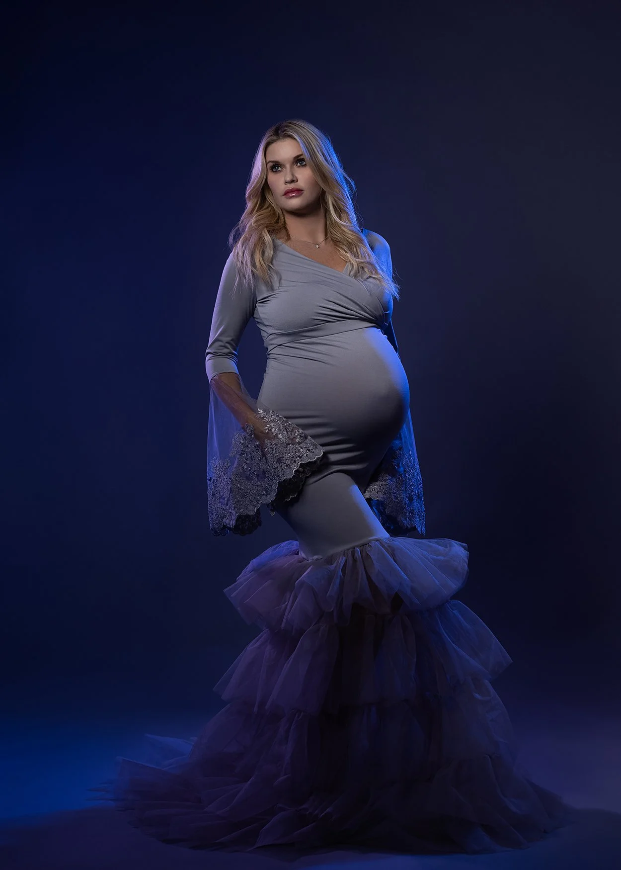 A pregnant woman in a stylish gray dress with lace and ruffles posing against a dark background with blue lighting.
