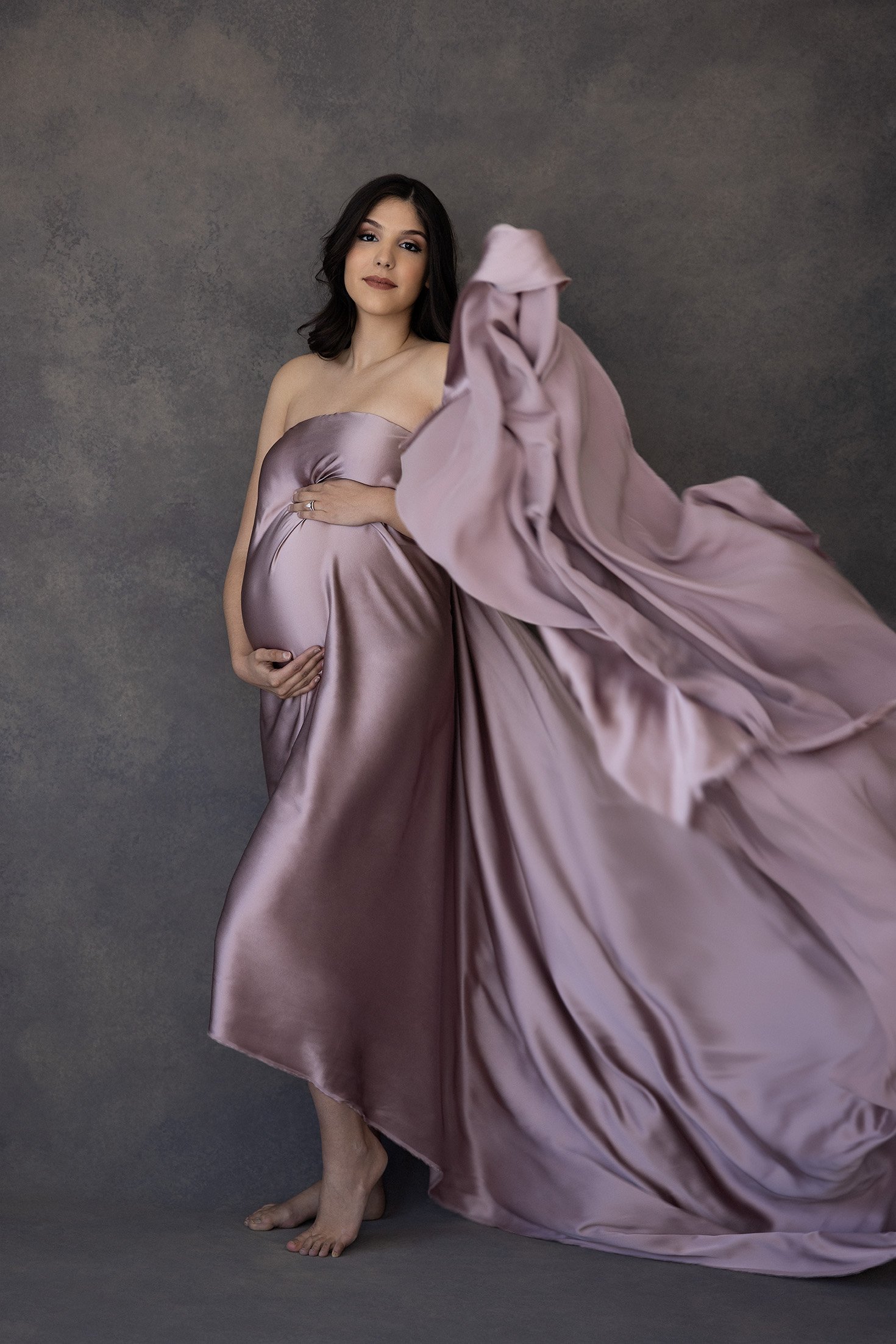 Pregnant woman in a metallic gown with a flowing train, posing barefoot against a gray backdrop.