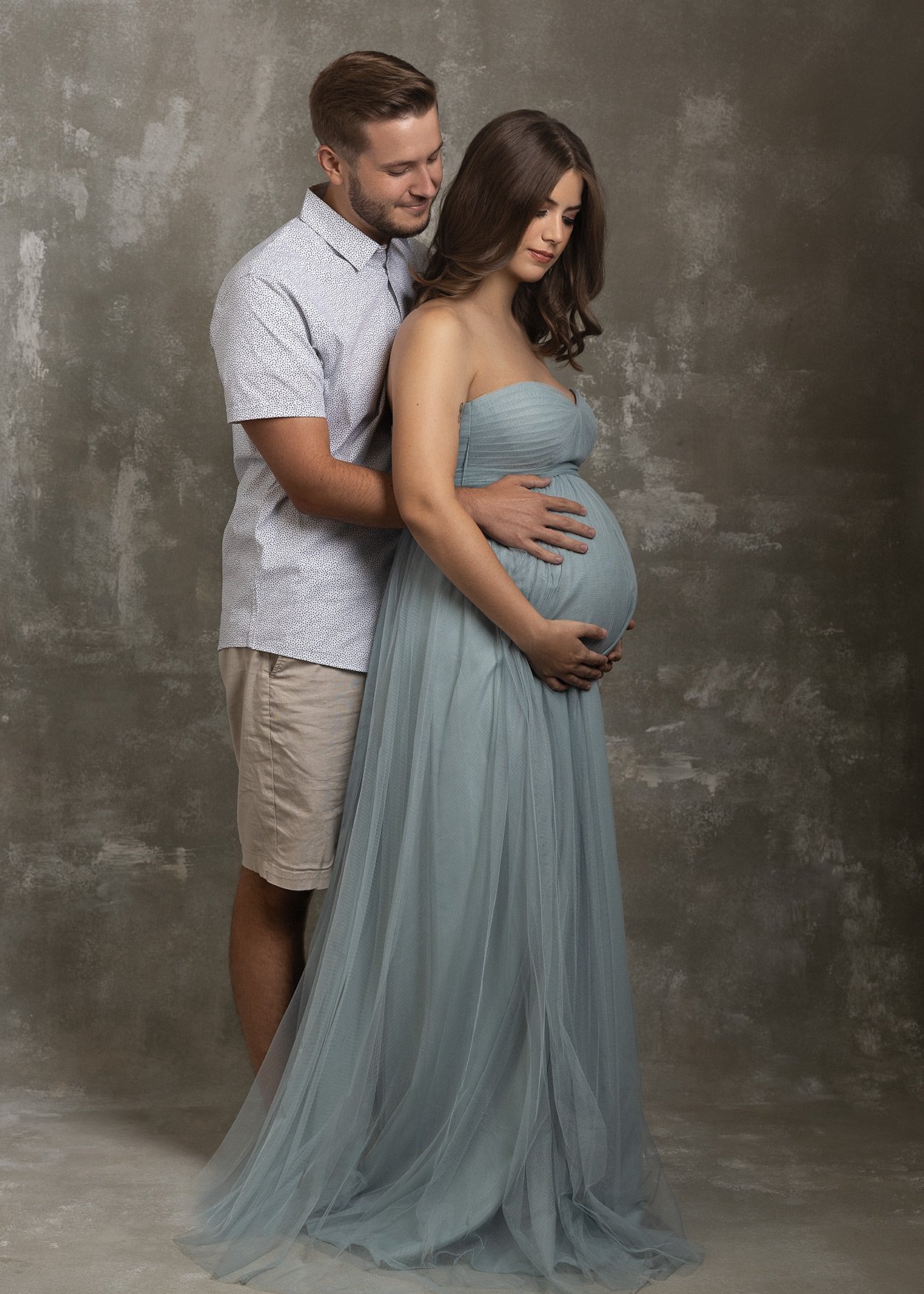 A pregnant woman in a strapless blue dress with wavy brown hair, cradling her belly, standing in front of a man in a white patterned shirt and beige shorts, with both looking down at her belly. They are posing against a textured gray background.
