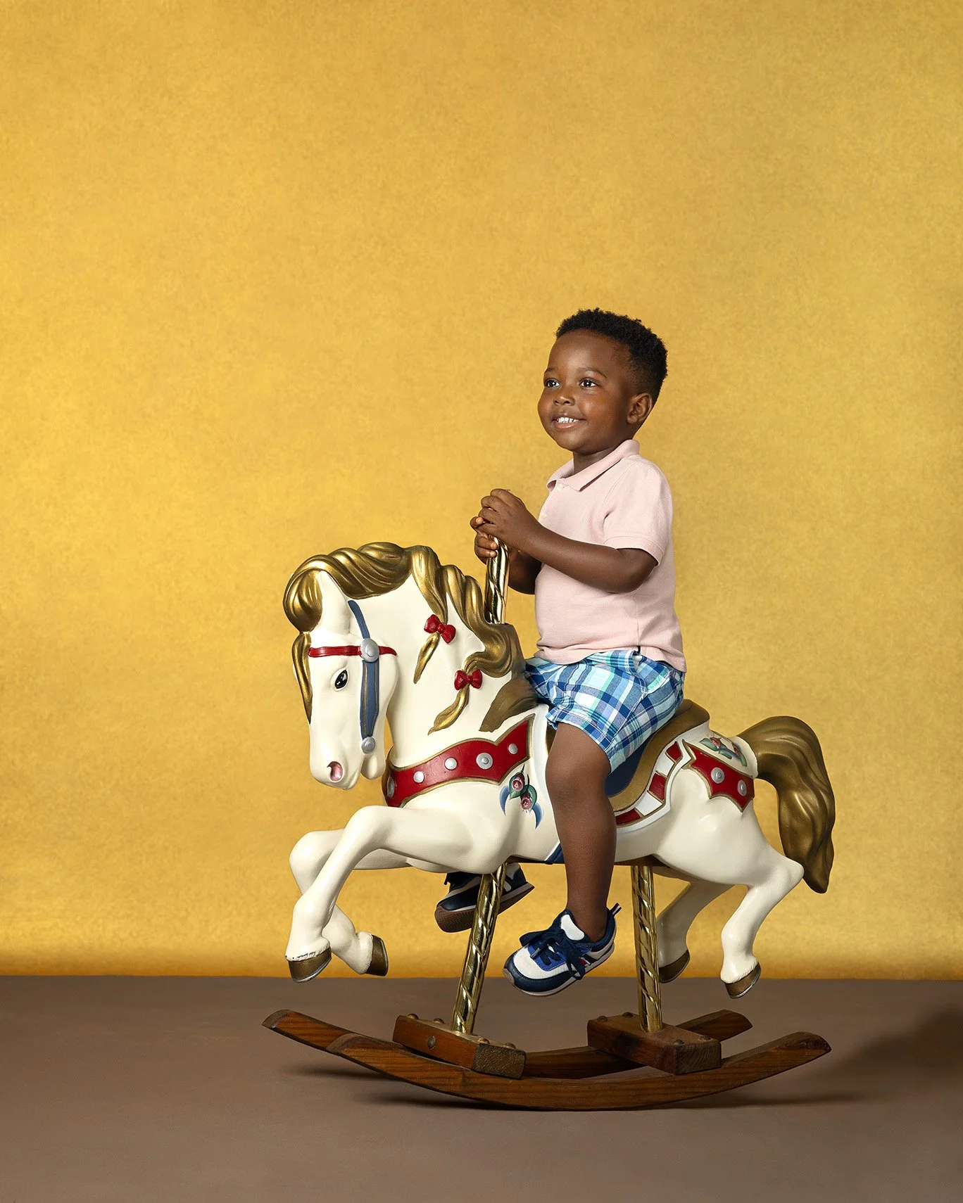 A young boy sitting on a white carousel rocking horse with gold and red details, against a yellow background.