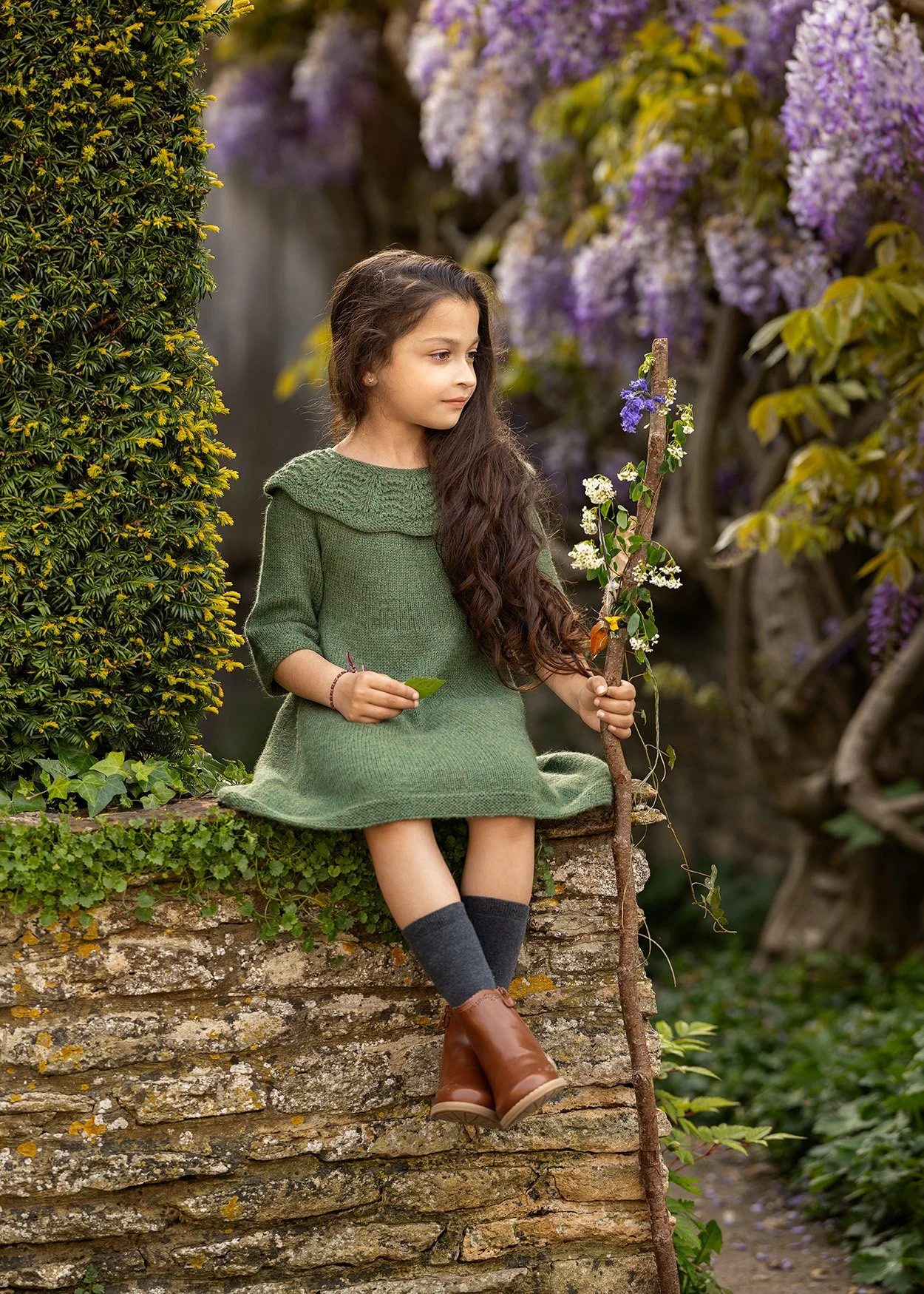 A young girl with long, wavy brown hair is sitting on a stone ledge in a garden. She is wearing a green sweater dress, gray knee-high socks, and brown boots. She holds a stick with flowering plants and a leaf in her hand and gazes thoughtfully to the side. Purple and white flowers bloom in the background.