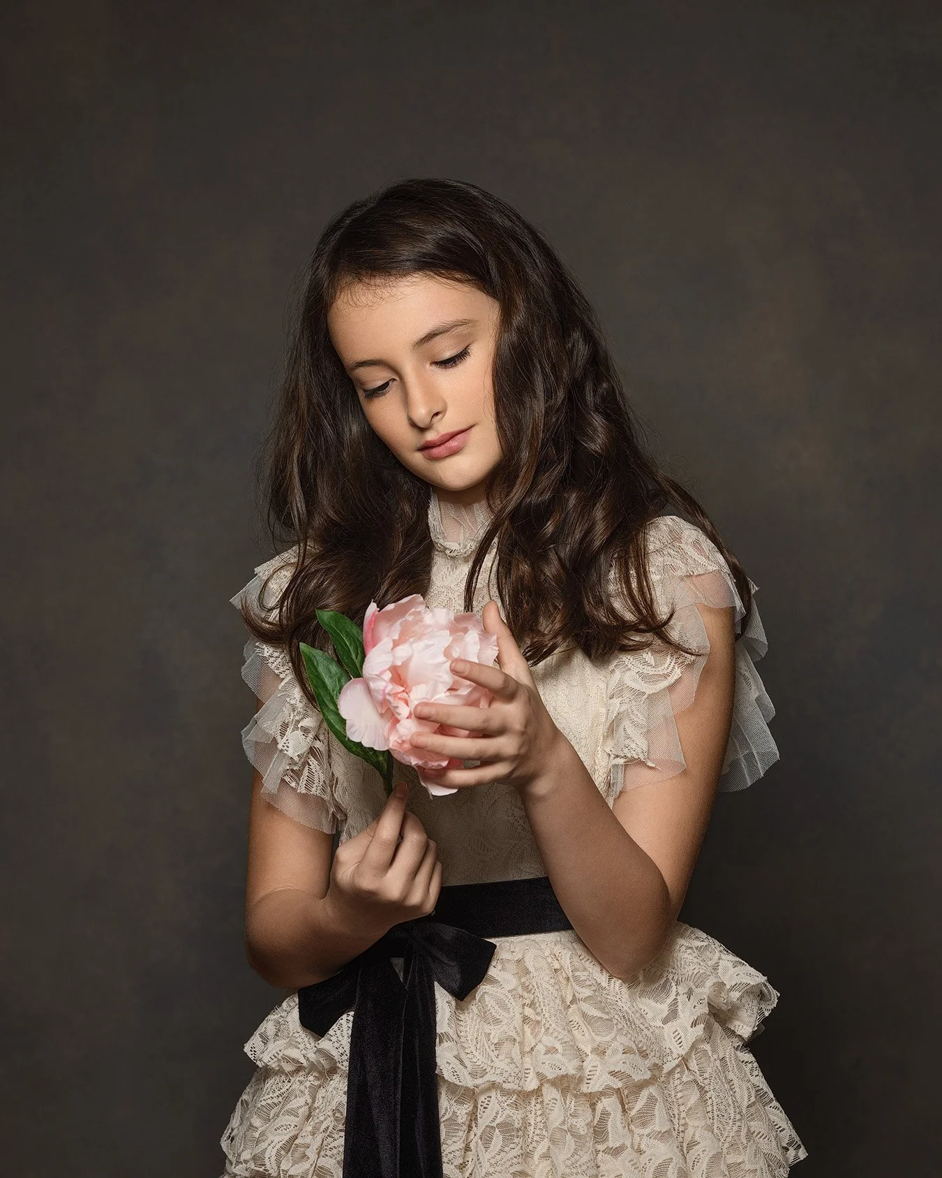 A young girl with long brown hair wearing a lace dress, holding a large pink flower, looking at it softly.
