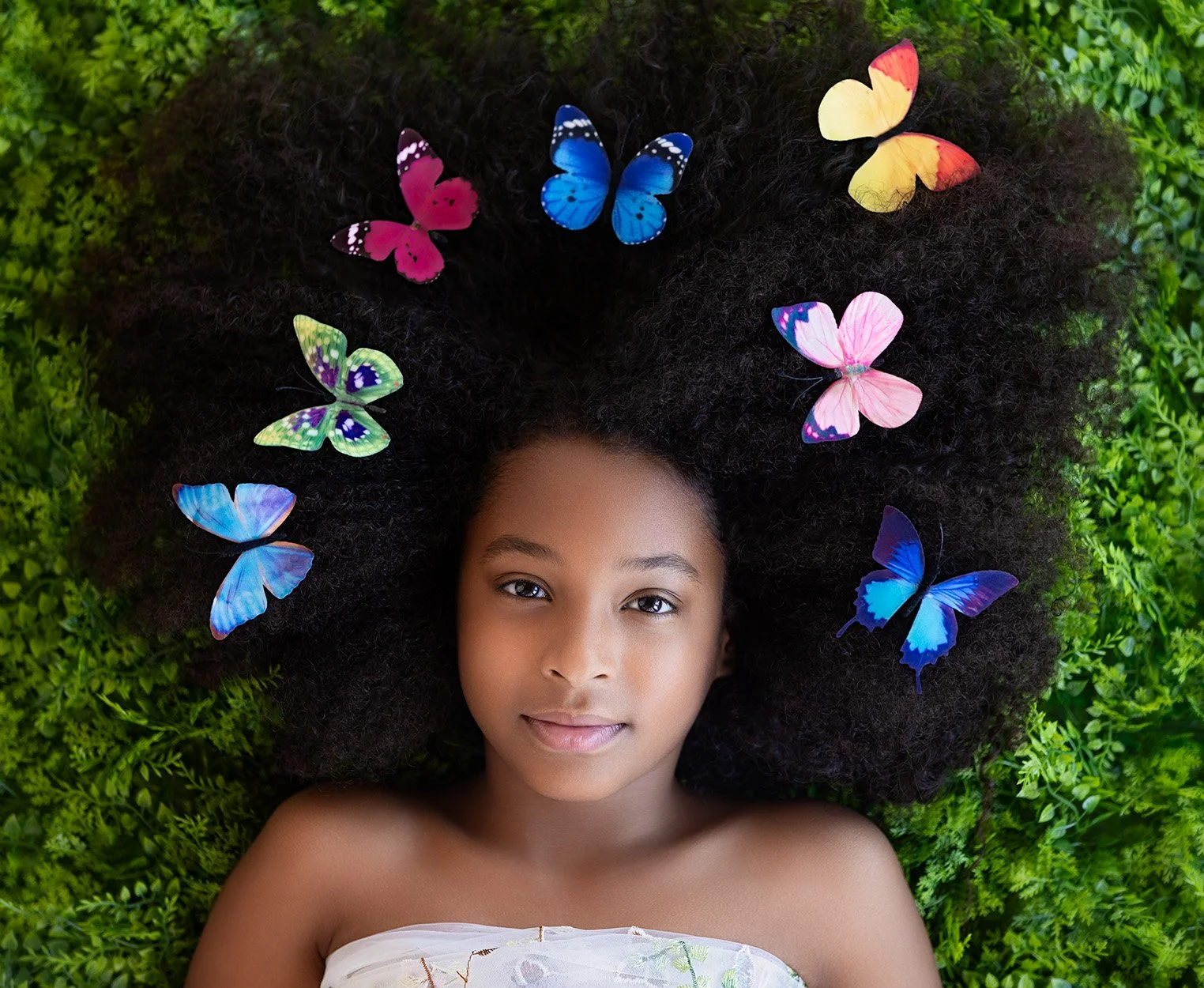 A girl lying on green grass with curly hair decorated with colorful butterflies.