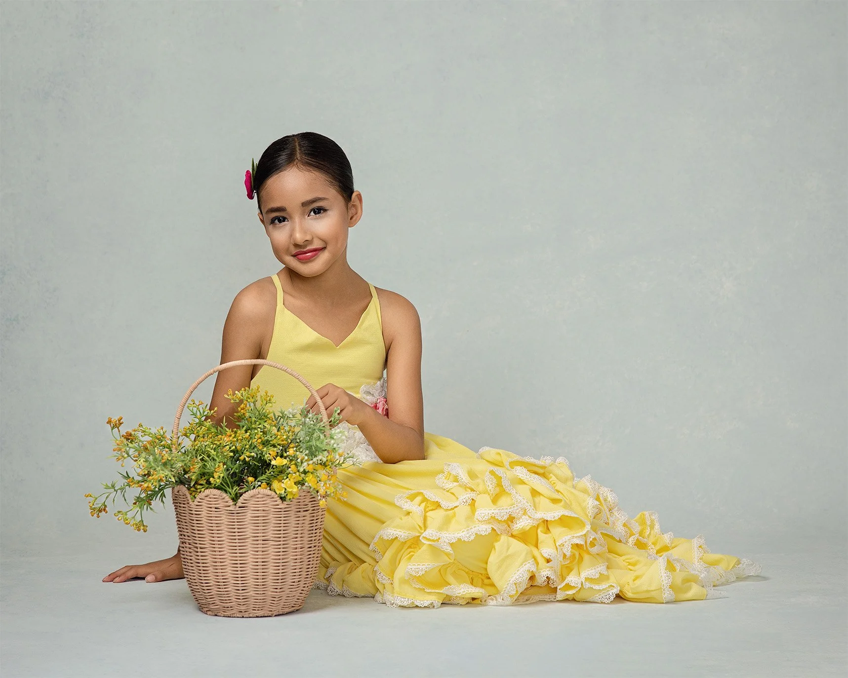 A young girl in a yellow dress sits on the floor next to a basket of yellow flowers, smiling at the camera.