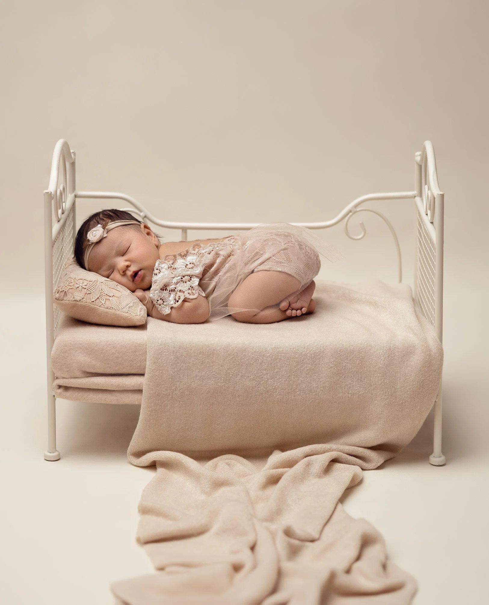 A young baby girl sleeping on a small beige bed with a matching pillow, dressed in lace clothing and wearing a headband with a flower, in a neutral background.