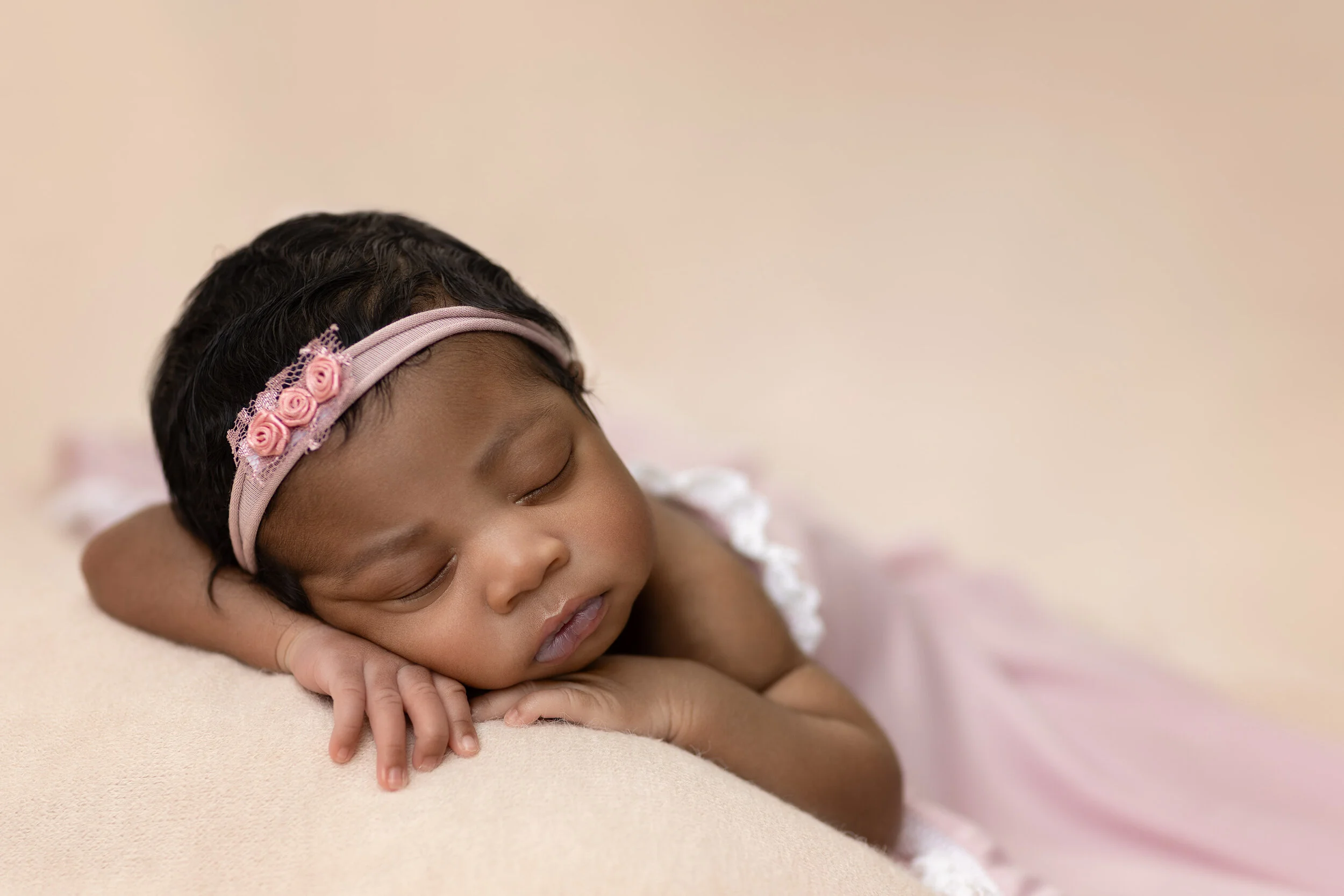 A young girl with dark hair, wearing a pink headband with pink rose details, peacefully sleeping on a soft surface with her head resting on her folded arms, dressed in a light pink outfit.