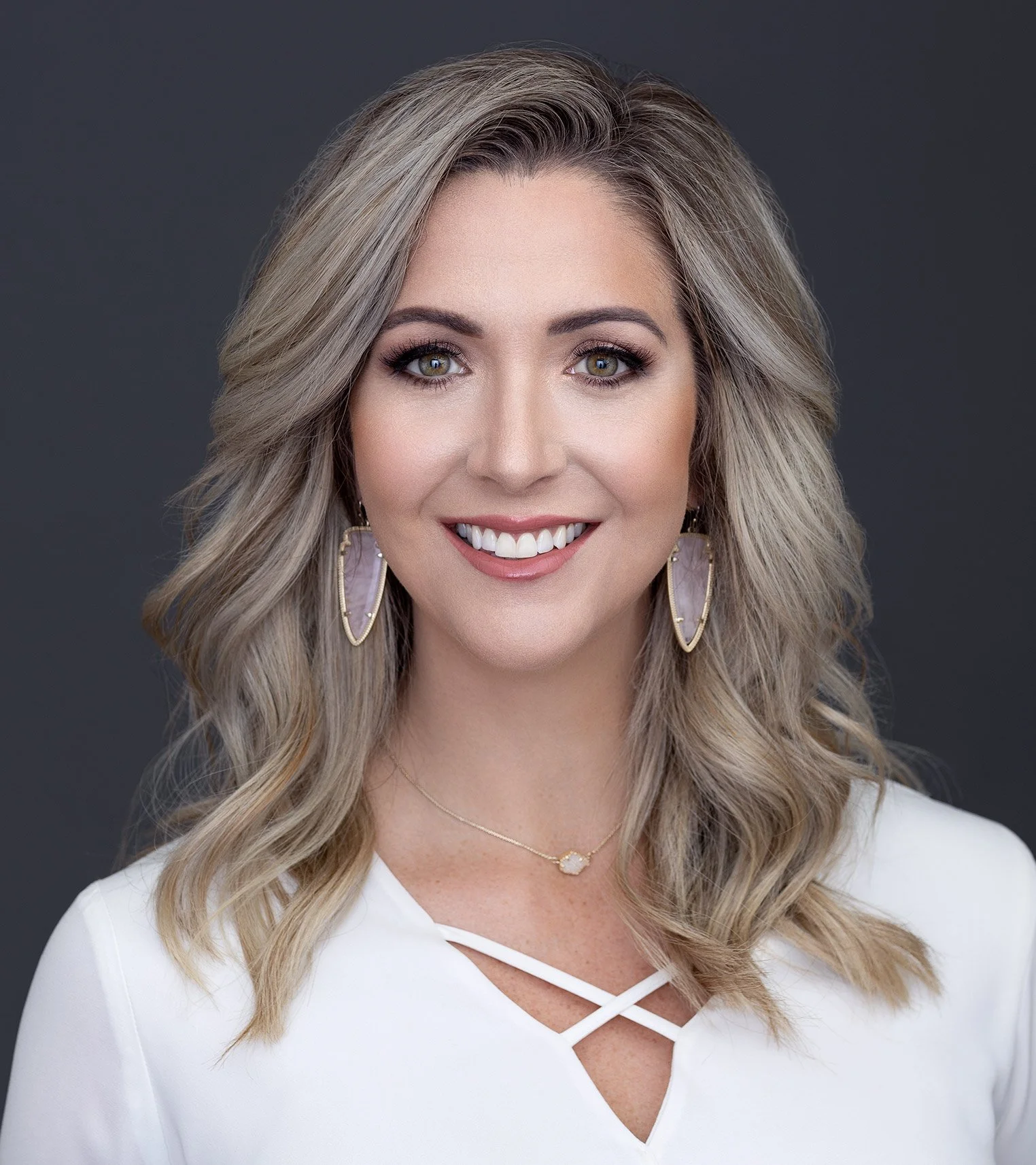 A smiling woman with wavy blonde hair, wearing a white top with crisscross detail, gold earrings, and a gold necklace, against a dark background.
