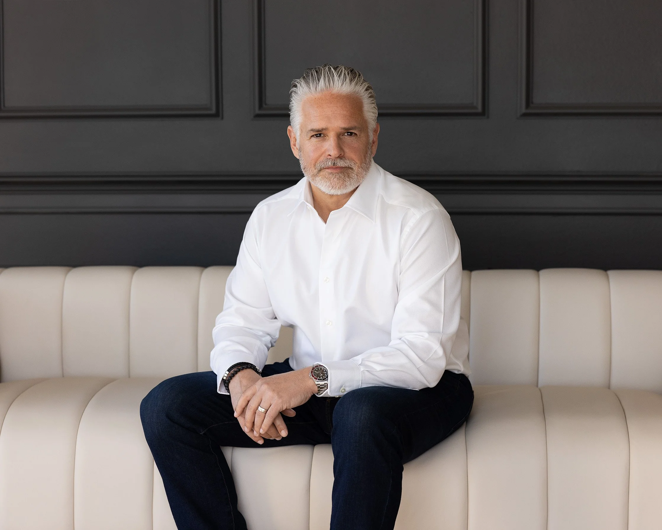 A middle-aged man with gray hair and beard, wearing a white dress shirt and black pants, sitting on a cream-colored sofa against a dark wall with decorative molding.