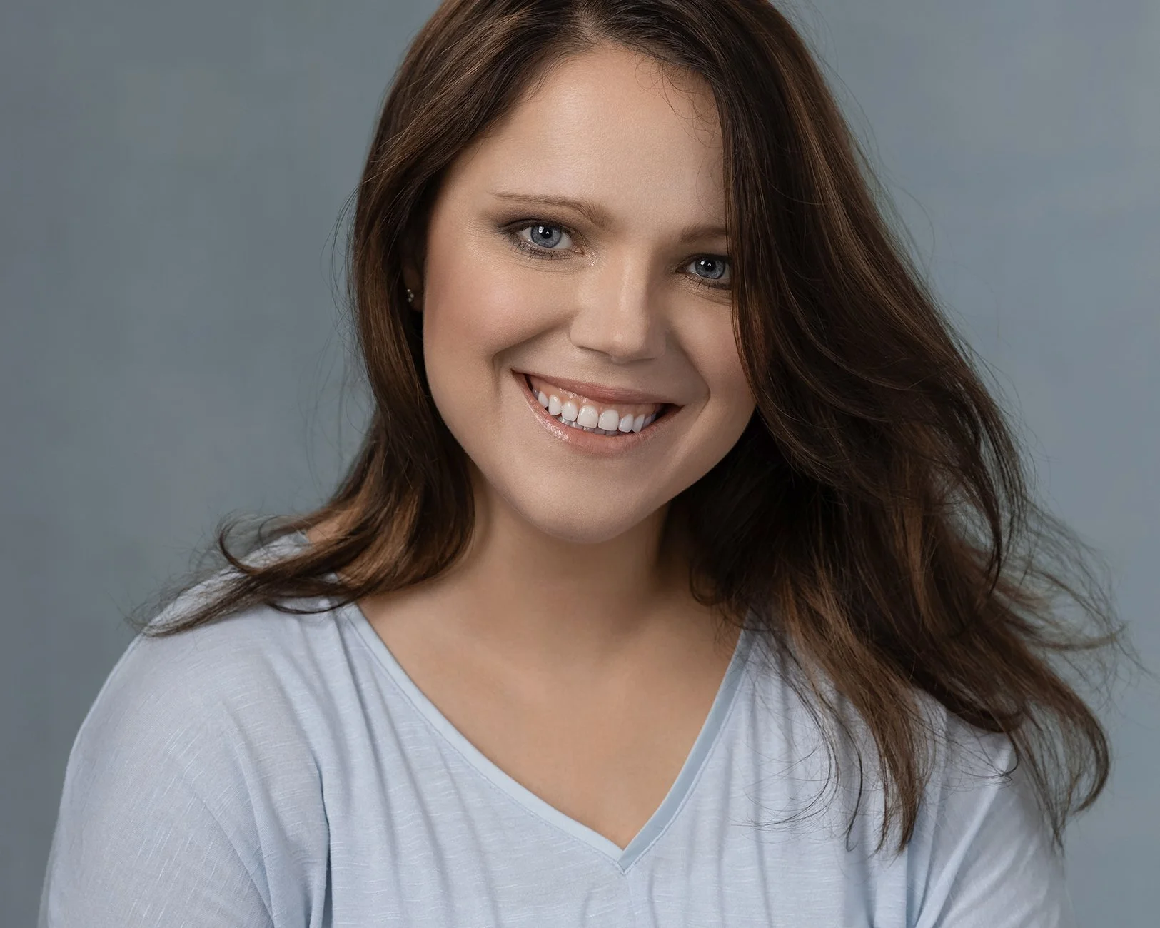 A woman with long brown hair, blue eyes, and a big smile, wearing a white top, against a gray background.