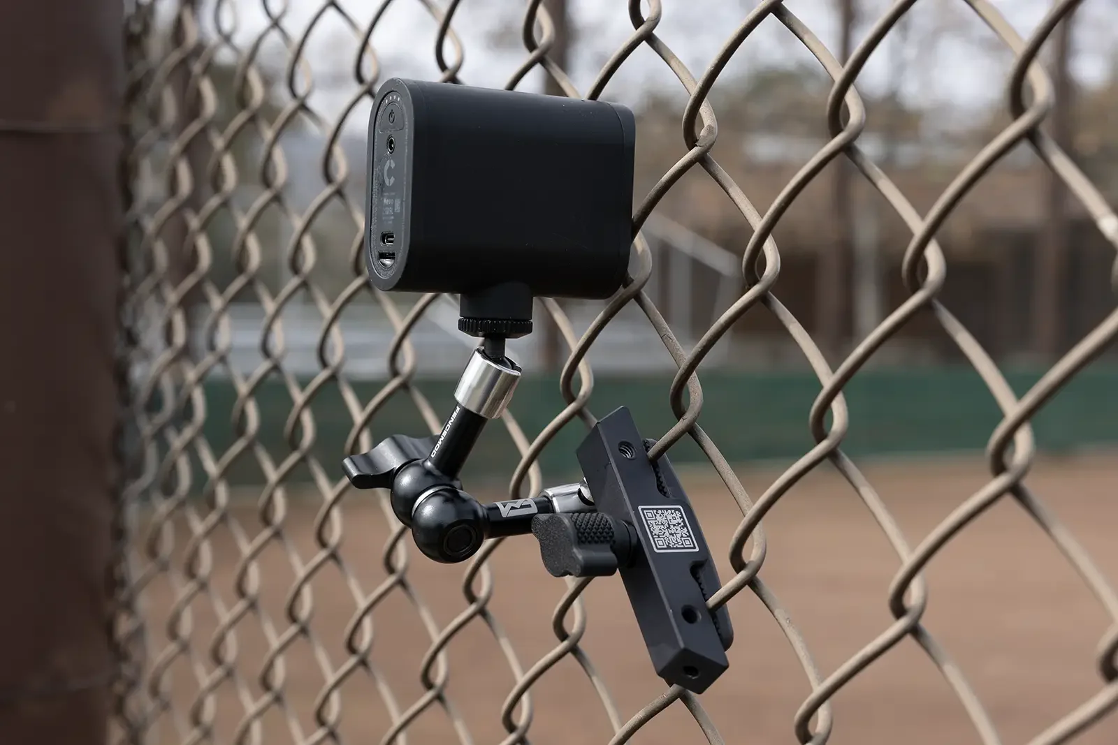 This image depicts a Mevo action camera mounted on a chain link fence using the Fence Mod Chain Link Fence Camera Mount.