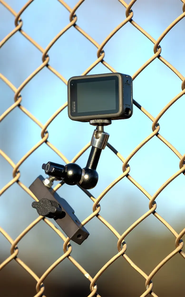 A GoPro camera mounted on a small tripod attached to a chain-link fence, with a blurred outdoor background.