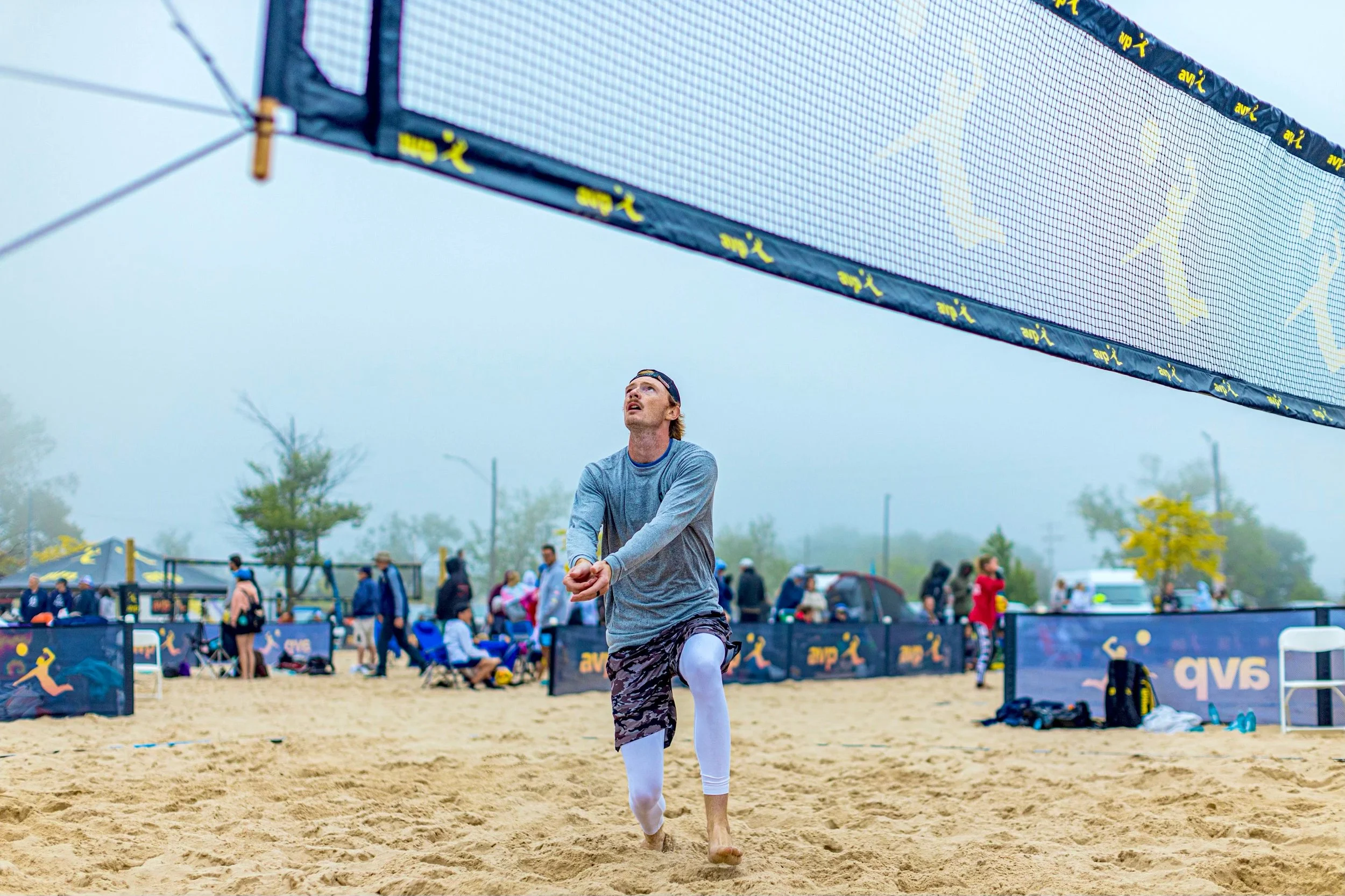 A male beach volleyball player preparing to hit a ball during a game on a sandy court with a foggy background and spectators watching.