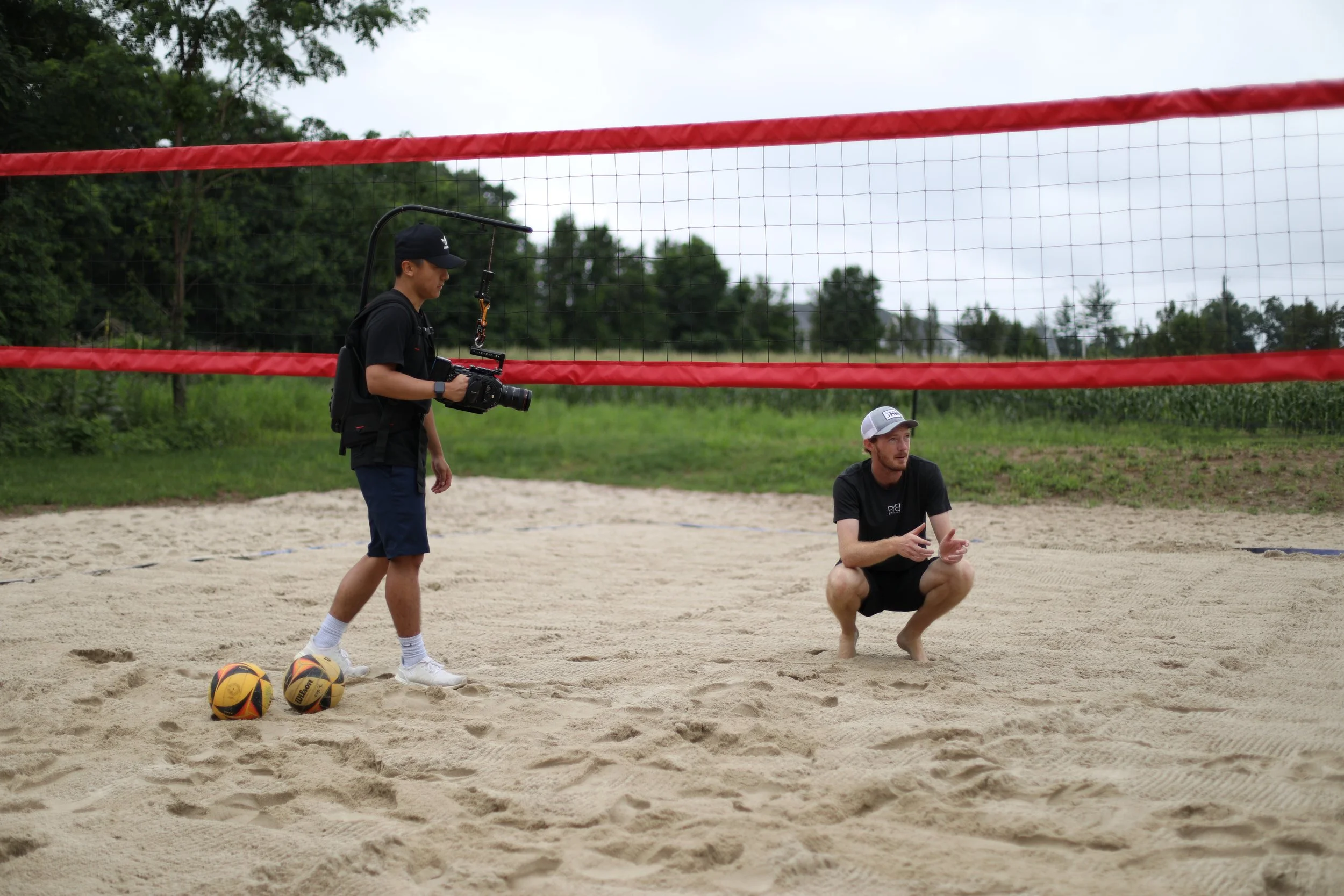 A person kneeling on a sand volleyball court, speaking, with a person standing nearby holding a camera, two volleyballs on the ground, and a volleyball net in the background.