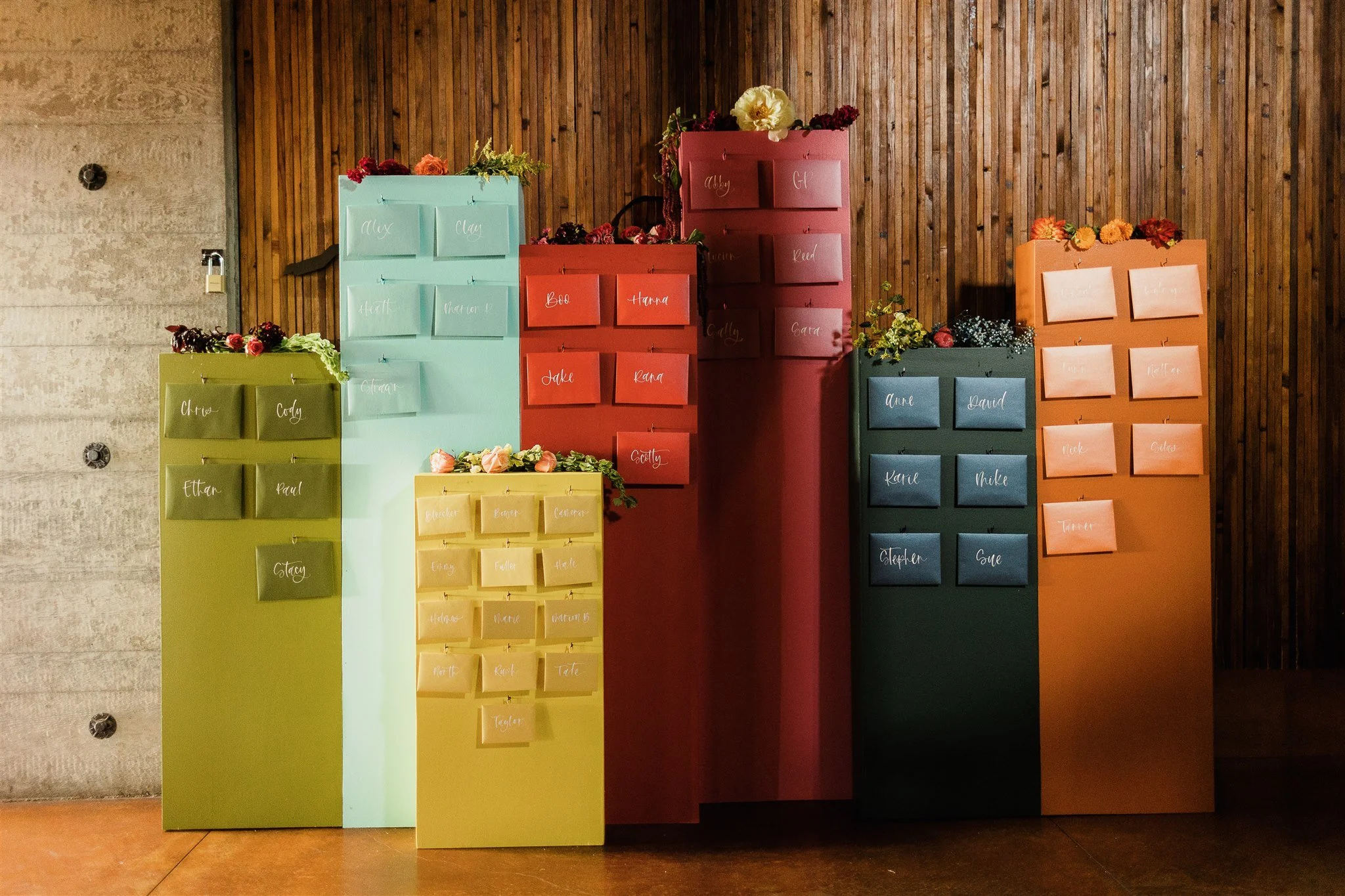 Colorful wooden lockers with handwritten names on each, decorated with flowers, arranged against a wooden wall, likely part of a wedding or event seating chart display.