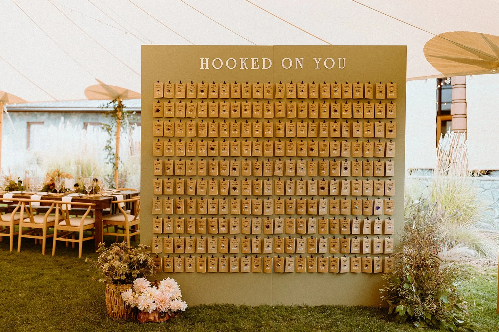A large display board with the words 'Hooked on You' at the top, filled with small hanging notes or tags, inside a decorated event tent with tables, chairs, and floral arrangements.