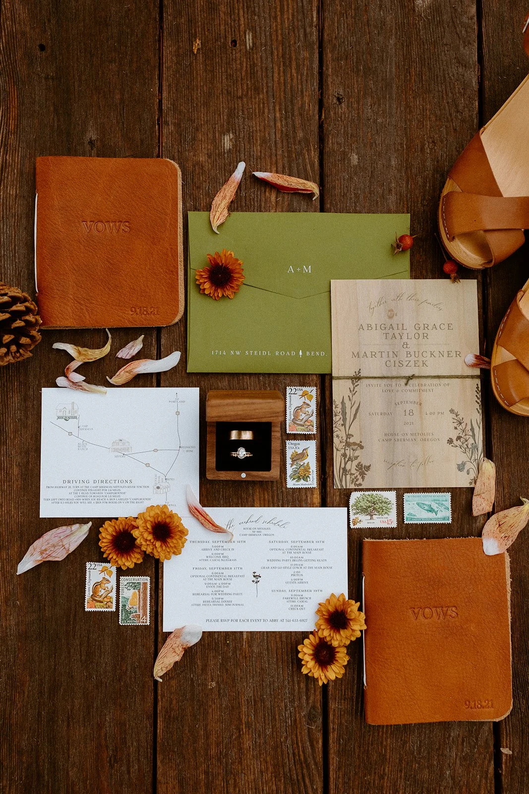 Wedding invitation and details, rings, postage stamps, and flowers arranged on a wooden surface for a wedding.