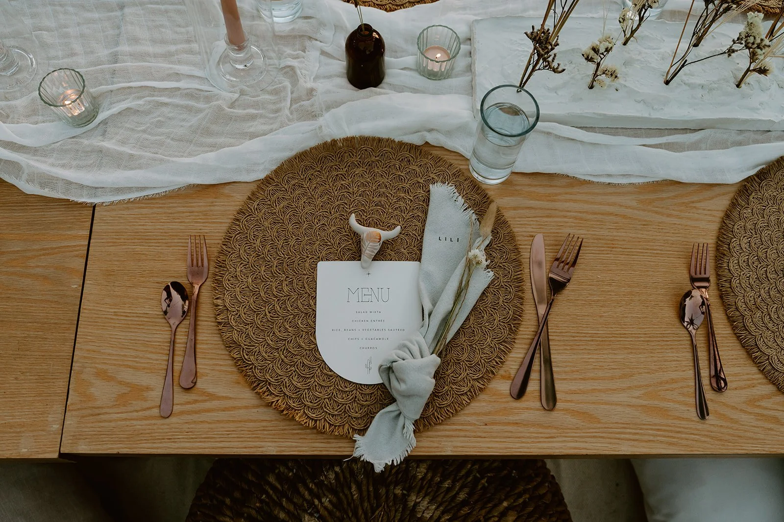 A table setting with a woven placemat, white menu card, cloth napkin knotted, two small cow-shaped decorations, and rose gold cutlery including a fork, knife, and spoon. There are glasses, a brown bottle with a flower, candles, and dried flowers in the background.