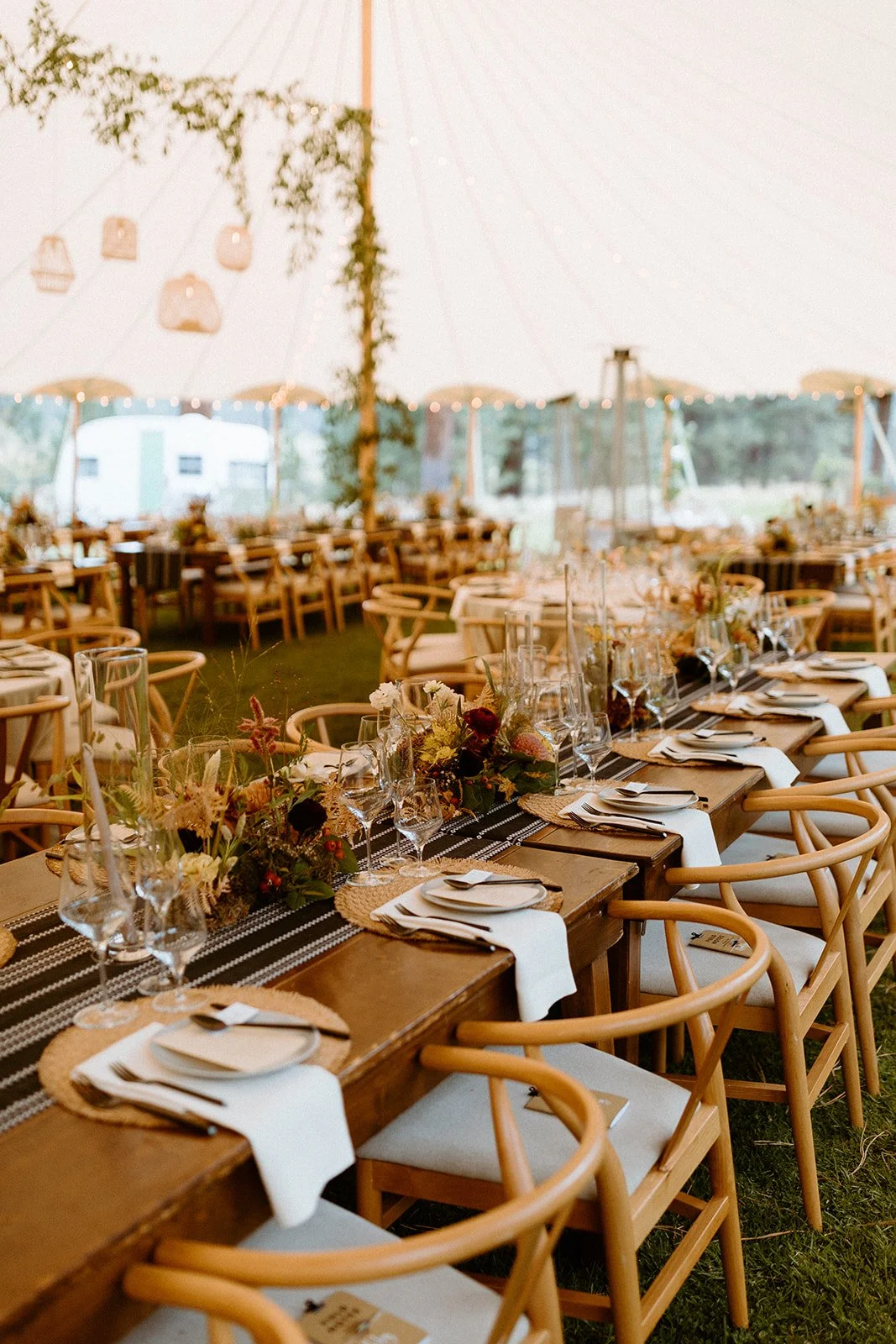 Wedding reception tent with a long wooden table decorated with floral centerpieces, glassware, plates, and napkins, surrounded by wooden chairs, under string lights and hanging lanterns.