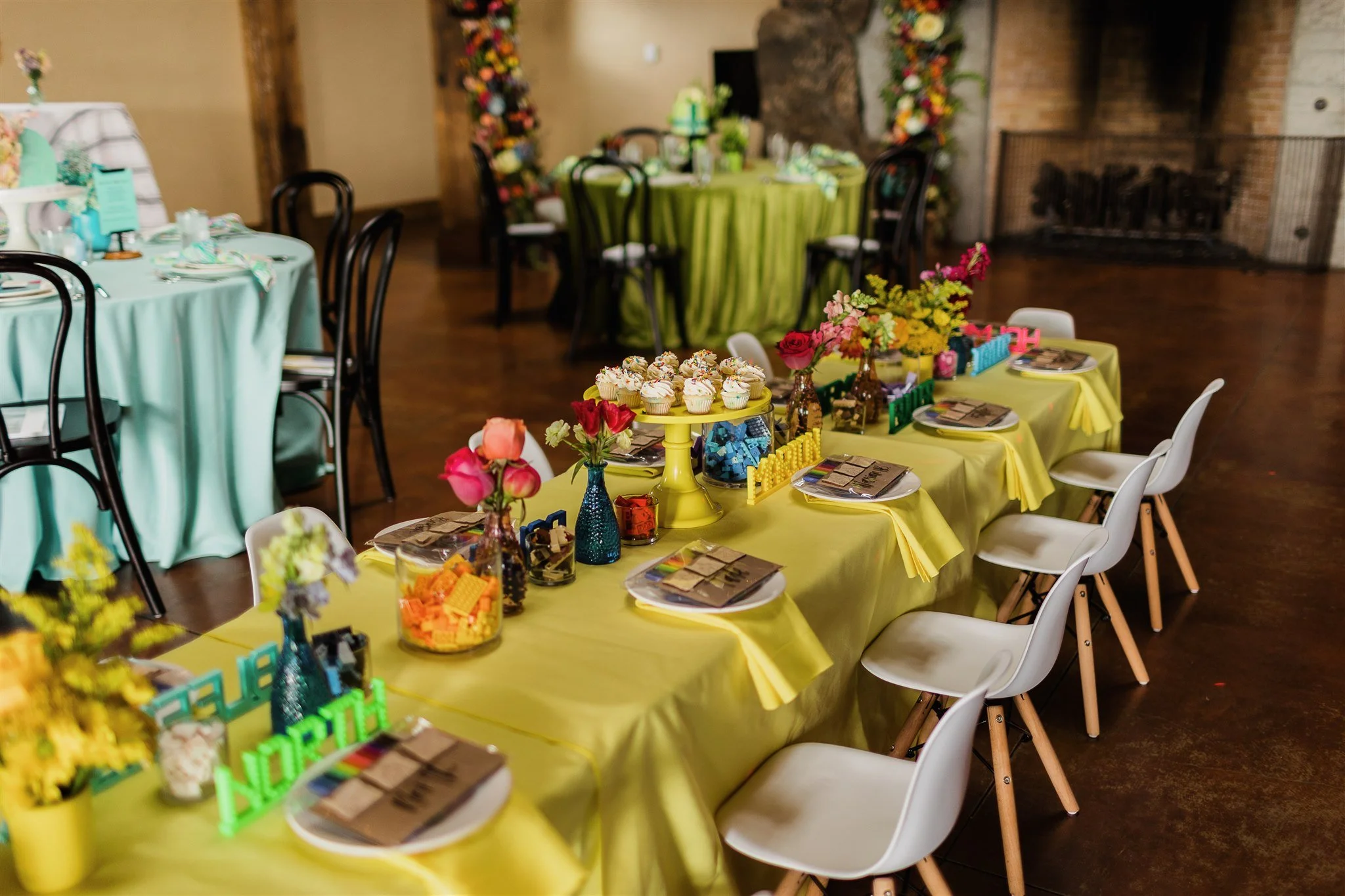 Colorful party table decorated with flowers, cupcakes, toys, and party favors. Multiple chairs are arranged around the table, with another setup of tables in the background. The setting appears to be indoors with a brick fireplace in the background.