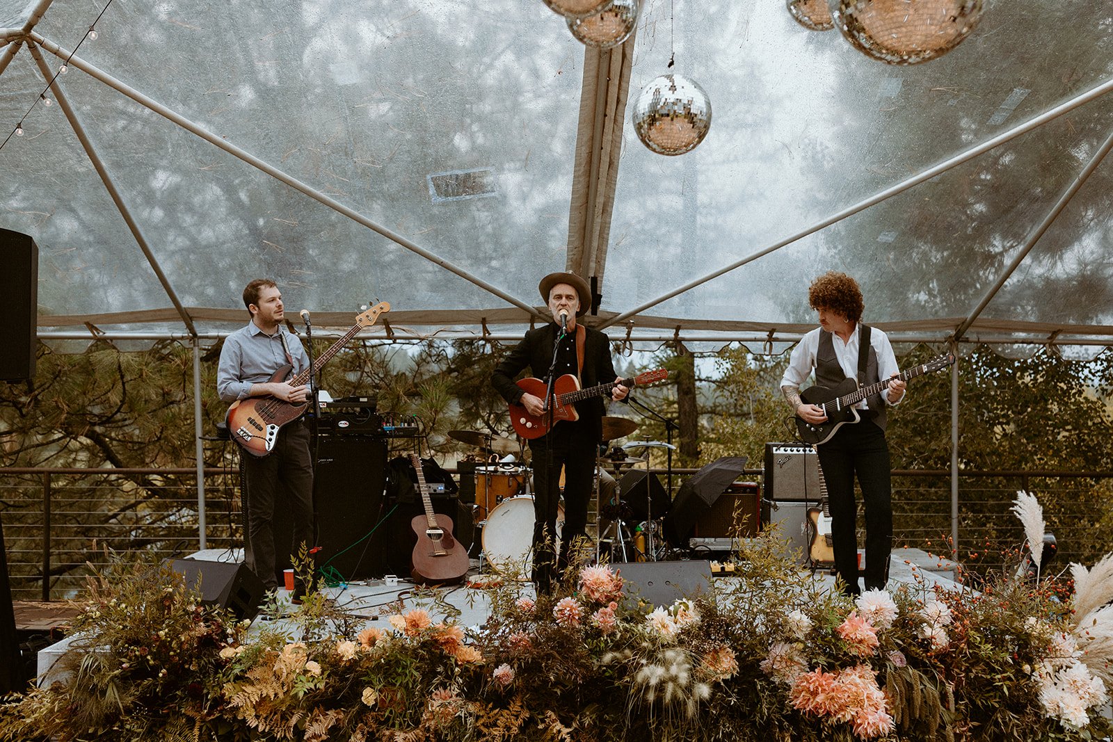 A band performing on a stage with three members playing guitars and a drummer in the background. The stage is decorated with flowers at the front and is set under a transparent tent with hanging disco balls. The outdoor setting features trees and cloudy sky.