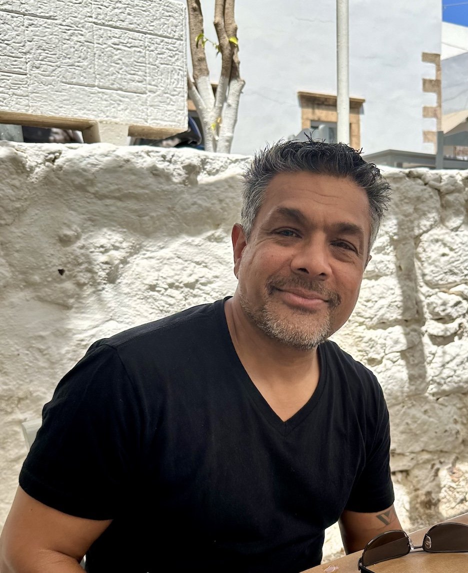 A man with short spiky dark hair with some gray, smiling, wearing a black t-shirt, sitting outdoors in front of a stone wall and trees.