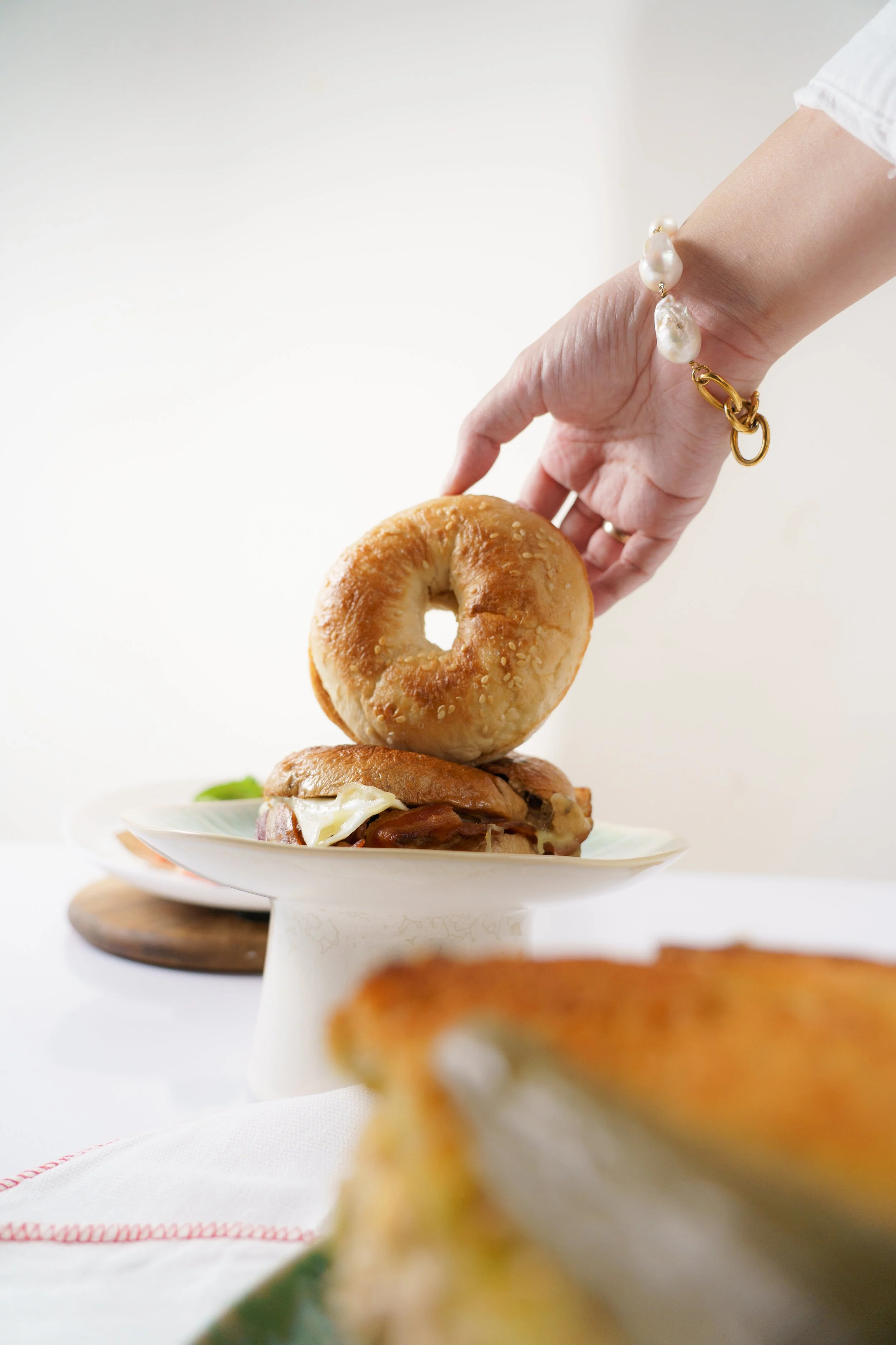 A hand with a pearl bracelet placing a bagel on a plate above a sandwich with melted cheese and ham.