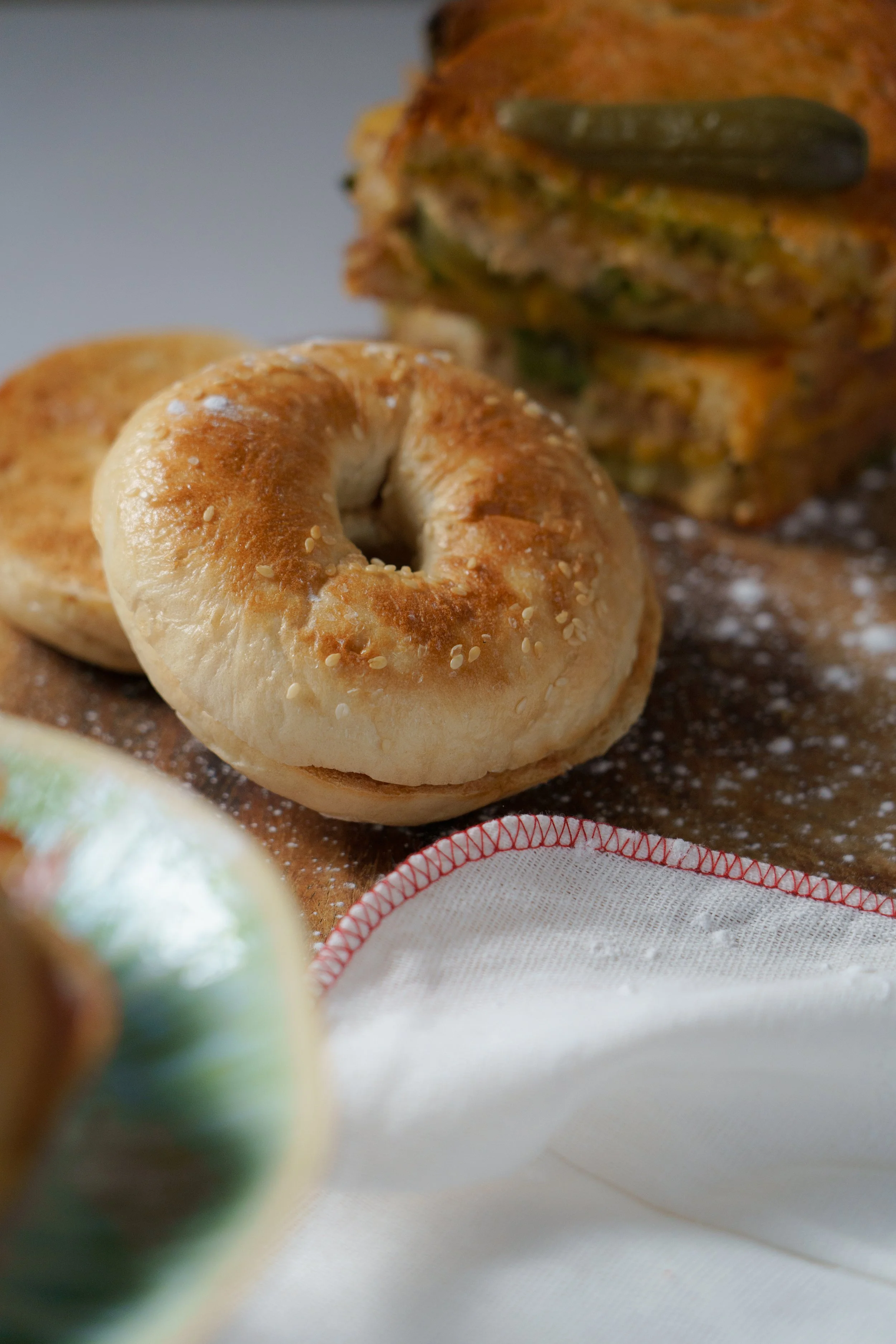 Close-up of a bagel with sesame seeds, a piece of layered casserole with zucchini and cheese in the background, and part of a decorated bowl and white cloth in the foreground.