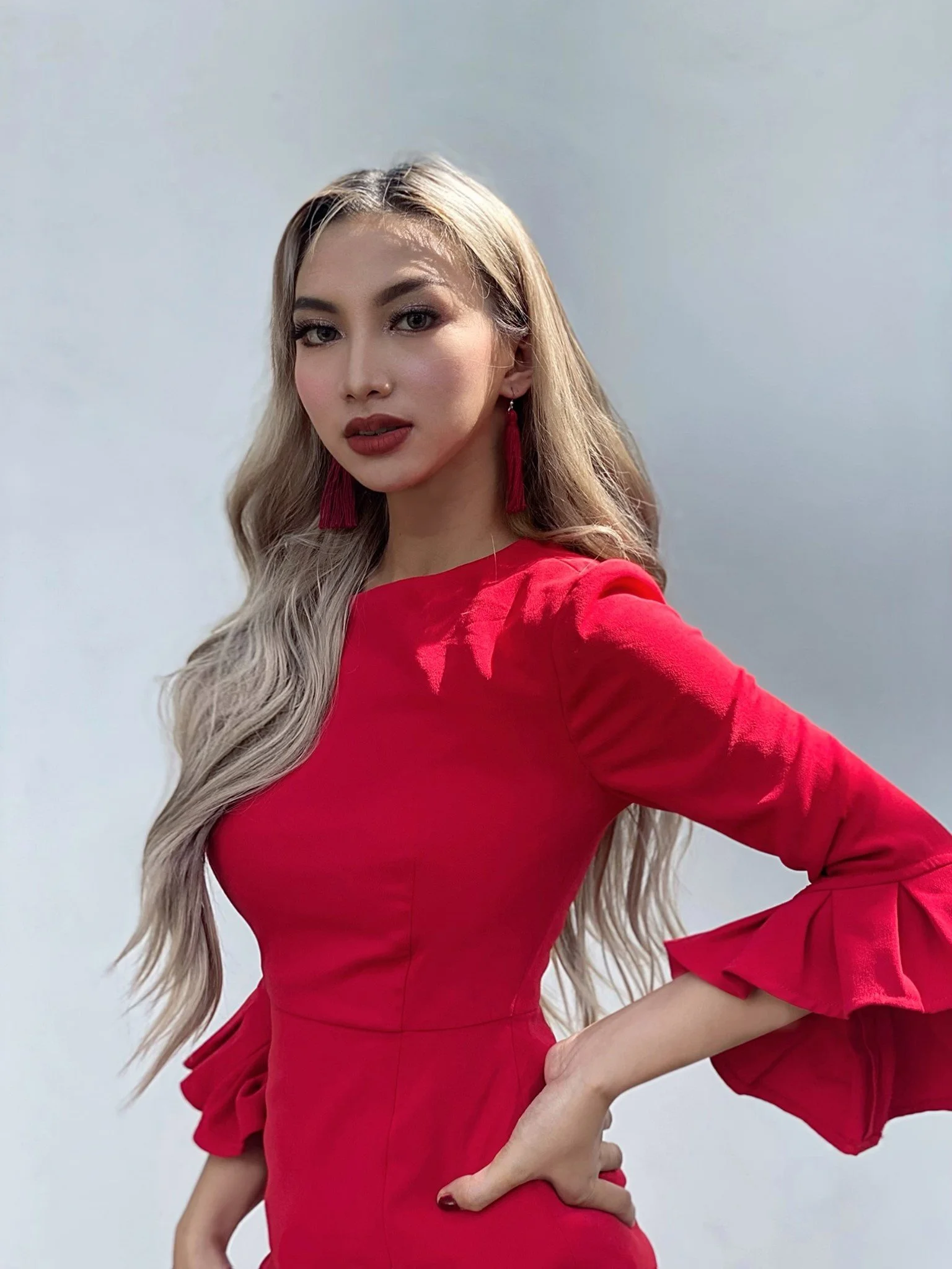 A woman with long, wavy blonde hair wearing a red dress with ruffled sleeves, red earrings, and makeup standing against a plain background.