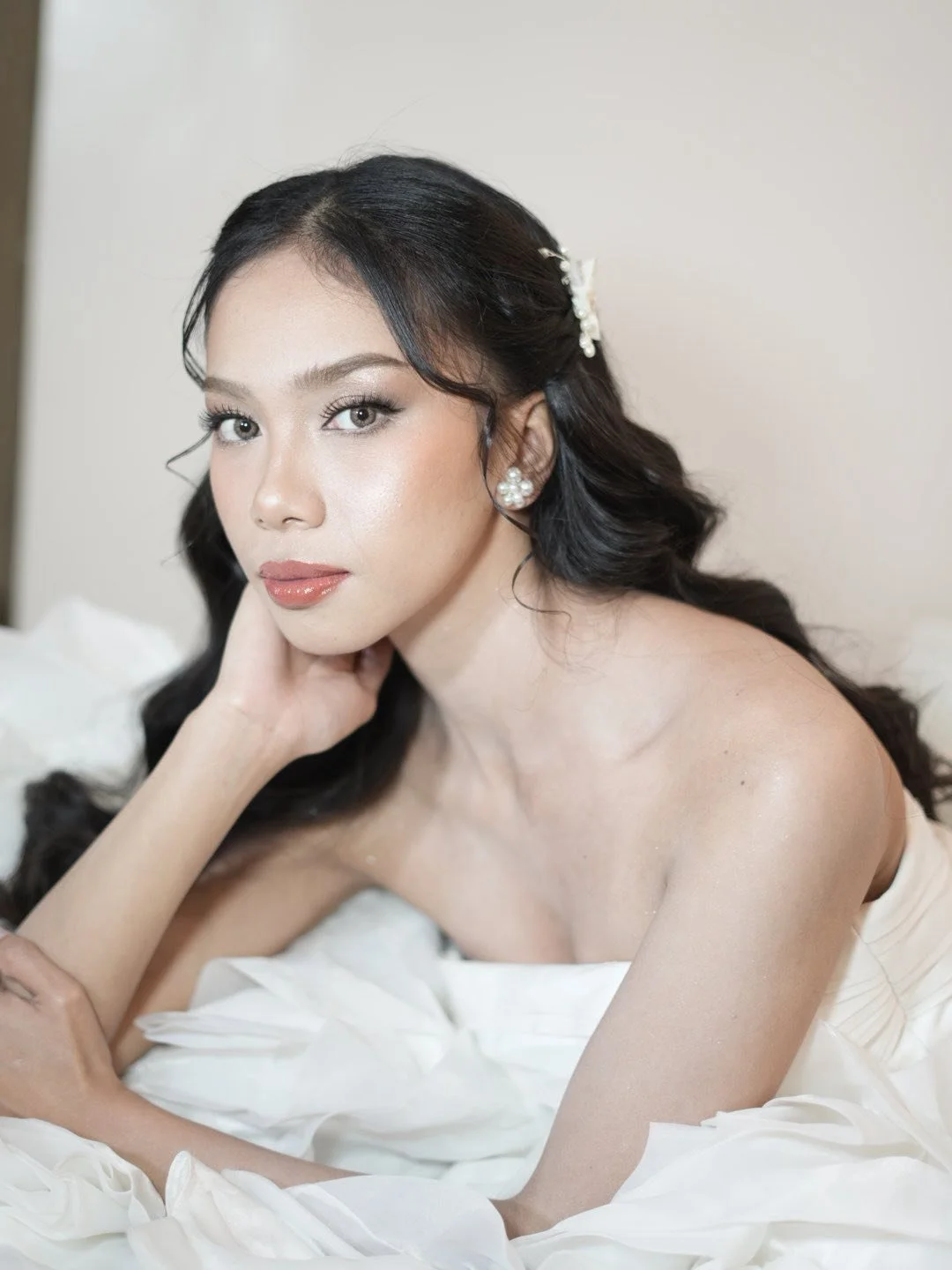 A woman with styled black hair, pearl earrings, and makeup, lying on a bed with white fabric.