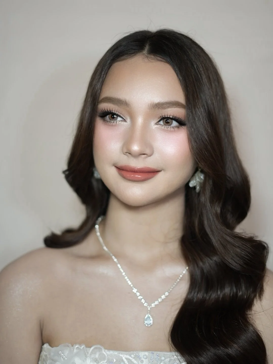 Portrait of a young woman with long dark hair styled in loose waves, wearing elegant jewelry including earrings and a necklace with a teardrop pendant, and a strapless dress with floral details.