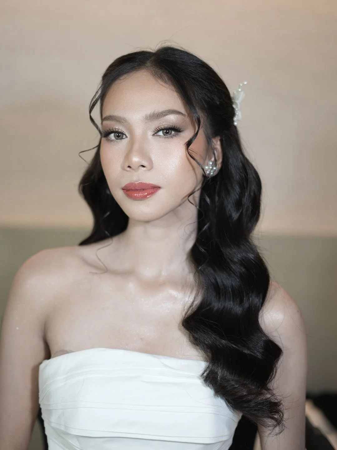 A woman with long black wavy hair wearing pearl earrings and a strapless white dress, looking at the camera with a neutral expression.