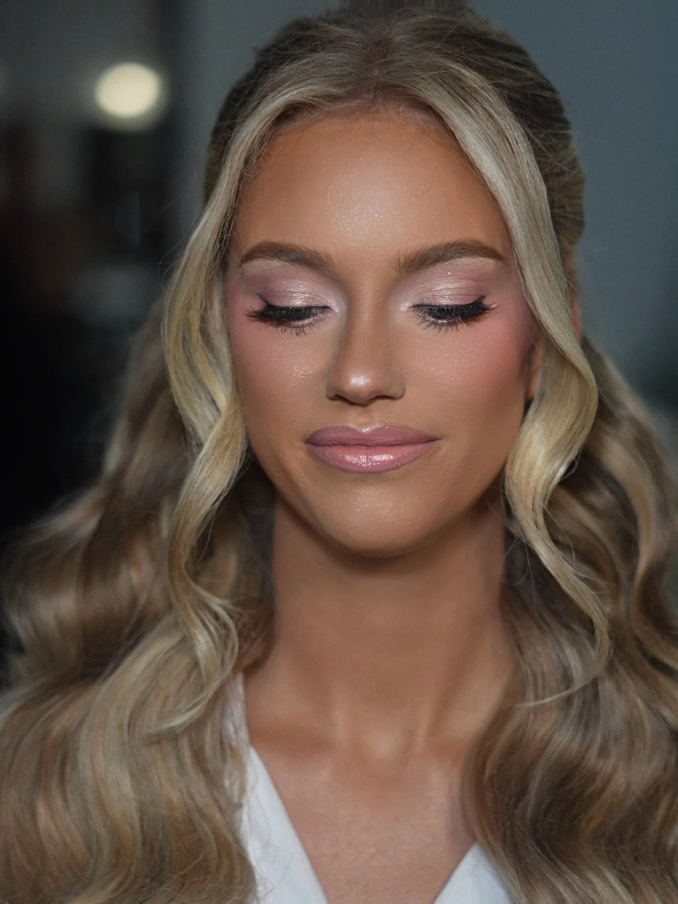 Close-up of a woman with long, wavy blonde hair, makeup with pink eyeshadow, eyeliner, and glossy lips, with eyes closed in a peaceful expression.
