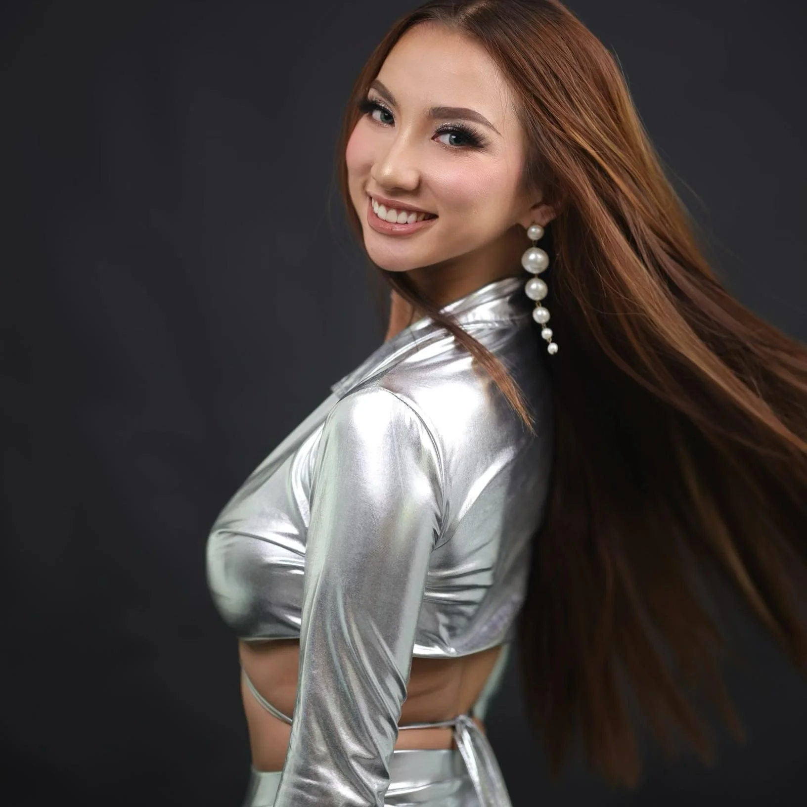 Young woman with long brown hair wearing a shiny silver outfit and large pearl earrings, smiling and posing against a dark background.