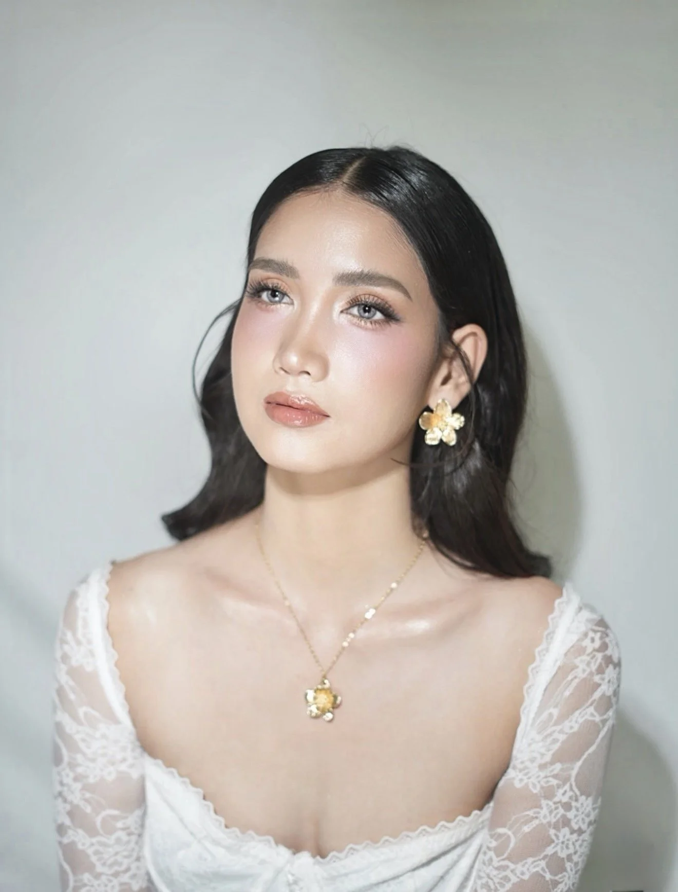 A young woman with dark hair and fair skin, wearing floral jewelry and a lace white dress, looking slightly to her right against a plain background.