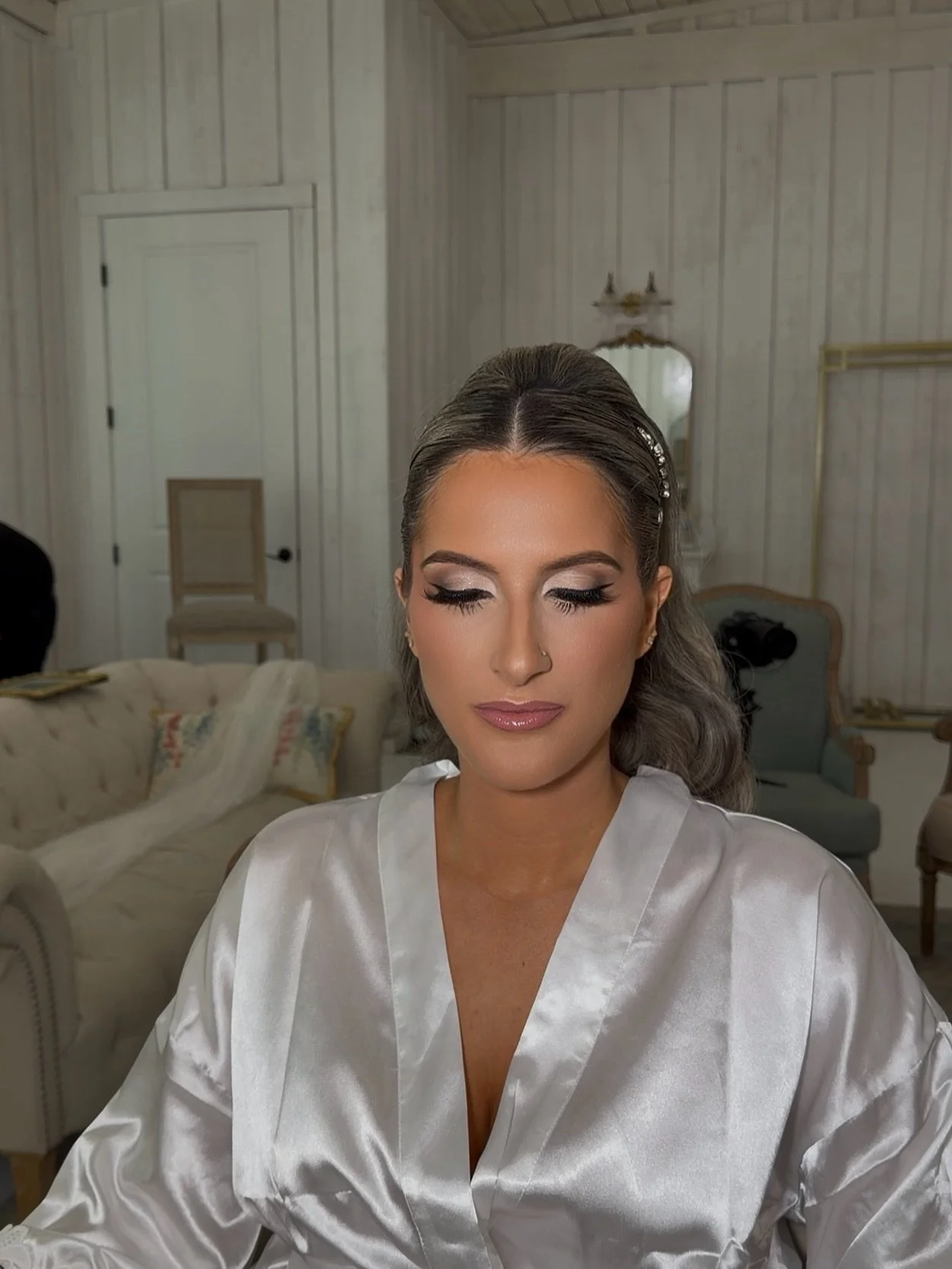 Woman with styled hair and makeup, wearing a satin robe, sitting in a room with vintage furniture and wood-paneled walls.